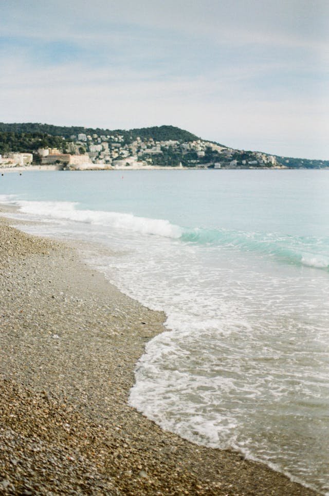 A pebble beach with gentle waves lapping onto the shore and a hillside town in the background under a partly cloudy sky.