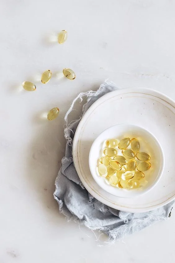 Vitamin capsules in a bowl on a white plate with a gray cloth underneath, some capsules spilled on a white surface.
