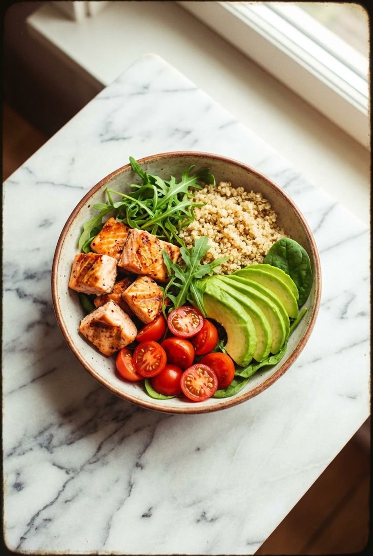 Bowl of grilled chicken, cherry tomatoes, sliced avocado, cooked quinoa, and greens on a marble surface near a window.