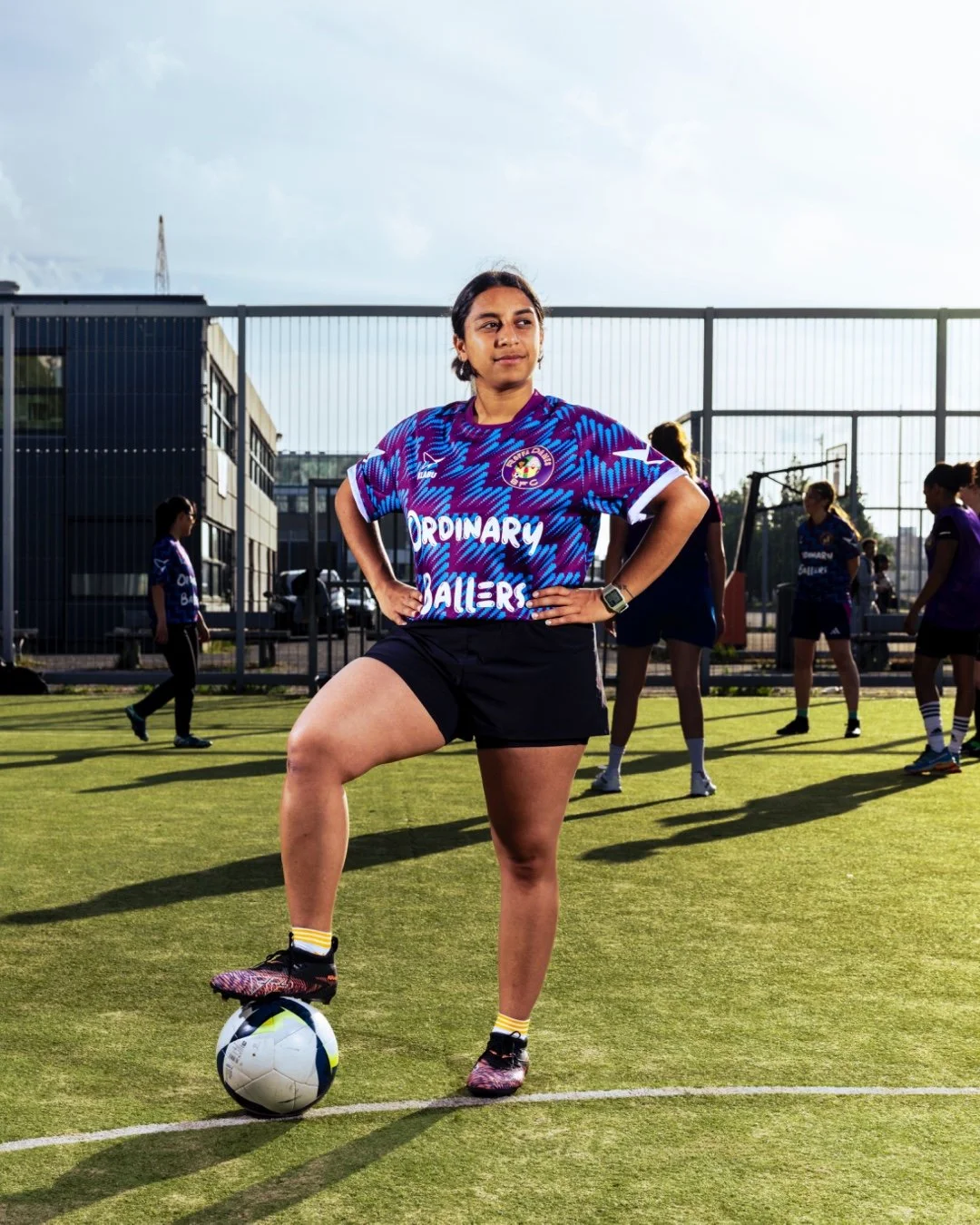 Young woman standing on a soccer field with one foot on a soccer ball, wearing a blue and purple sports jersey and black shorts, with other people in the background, during daytime.