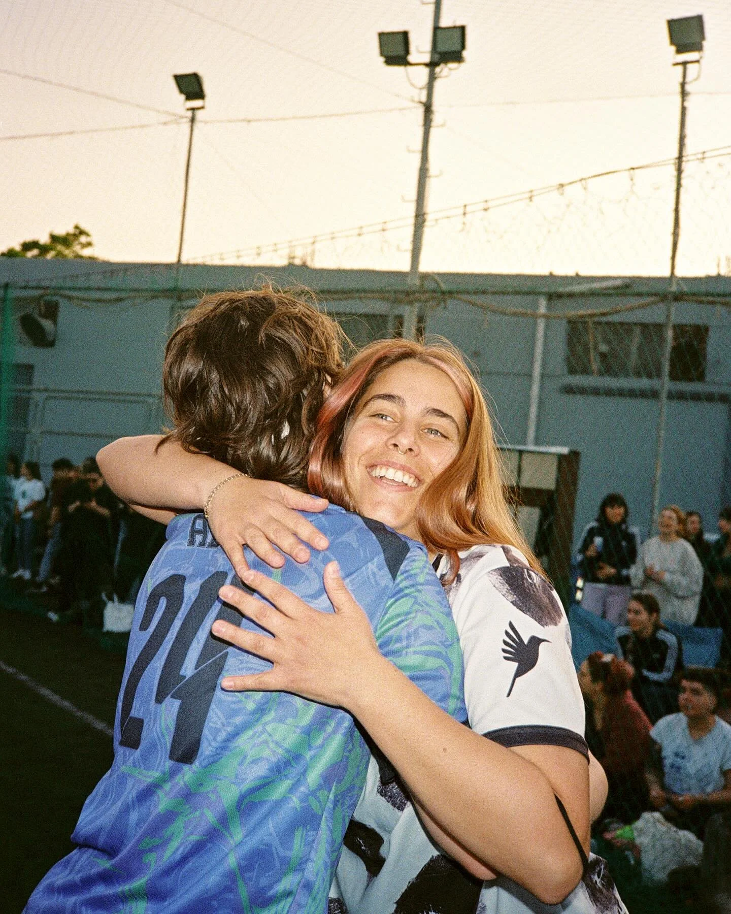 Two women hugging each other and smiling in an outdoor setting with a crowd of people in the background. The woman on the left has reddish hair and is wearing a blue sports jersey. The woman on the right has lighter hair and is wearing a white jersey with black designs.
