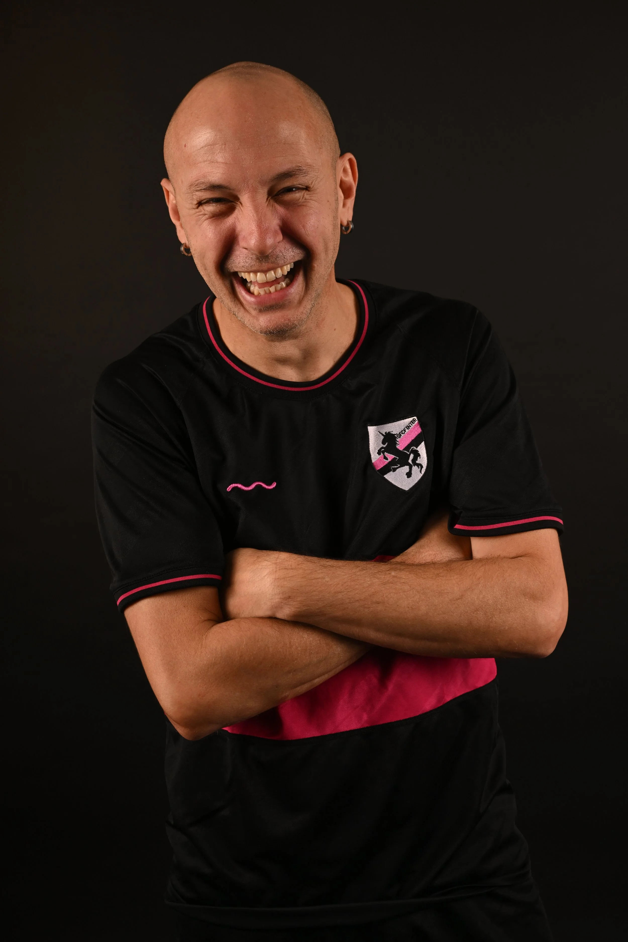 A smiling bald man with earrings, crossing his arms, wearing a black sports jersey with pink accents and a unicorn logo on the chest, against a dark background.
