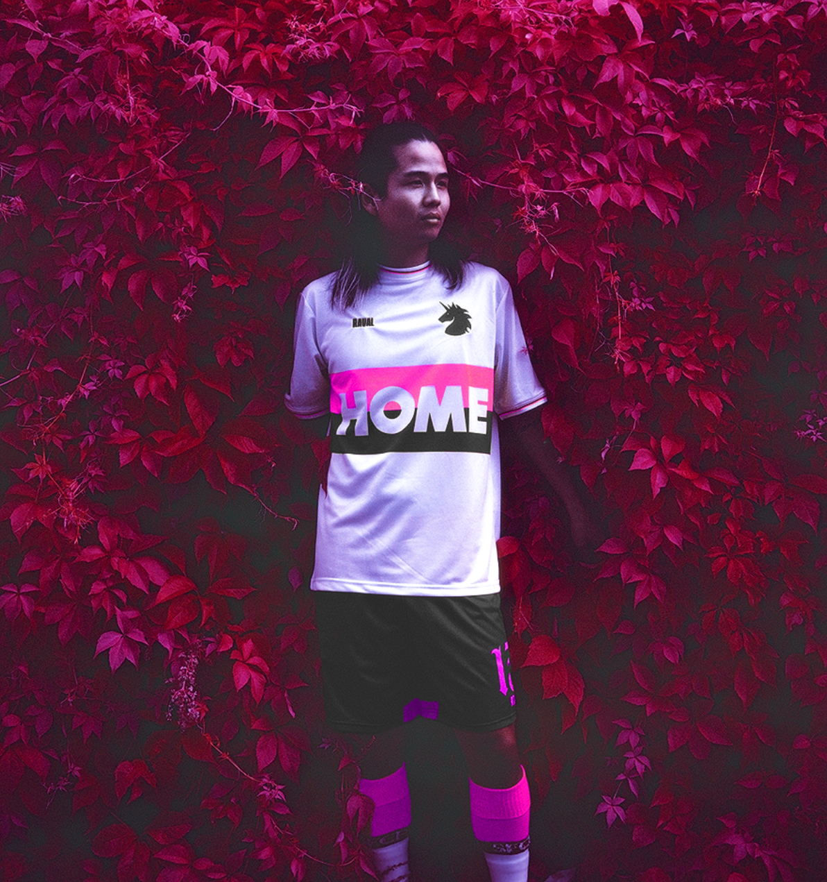 A young man in a white soccer jersey with pink, black, and purple accents, standing against a wall of vibrant red and purple leaves. The jersey has the word 'HOME' printed across the chest and a black unicorn silhouette logo on the left side. He has long dark hair and a serious expression.
