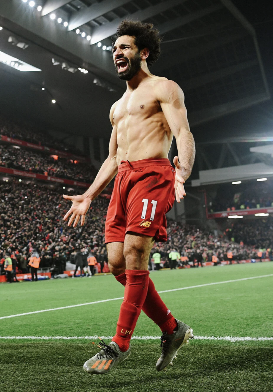 A shirtless male soccer player celebrating on the field in a stadium, wearing red shorts and socks, with a large crowd in the background.