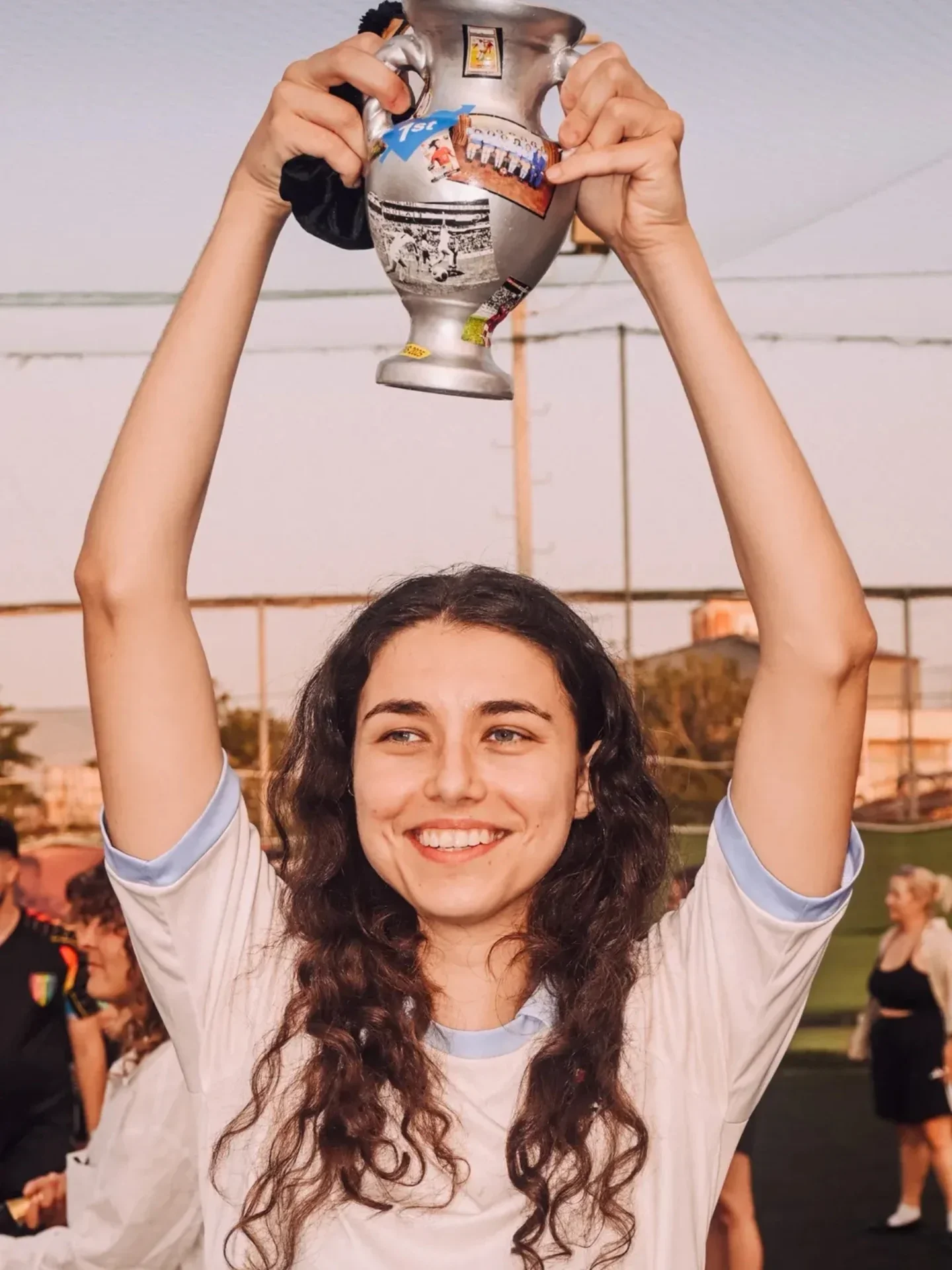 A young woman with long, curly brown hair smiling and holding a trophy above her head in an outdoor sports field, with other people in the background.