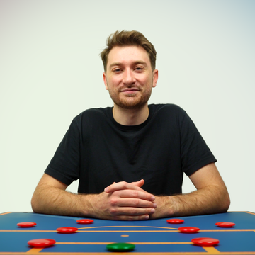 A young man sitting at a table with a blue surface and black lines, with red and one green game piece, smiling at the camera against a plain light-colored background.