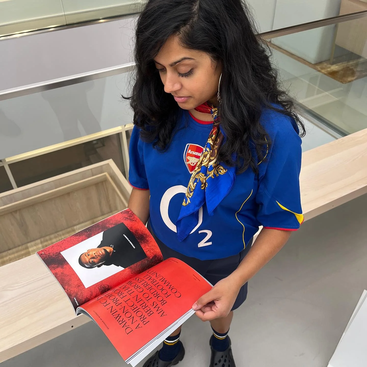 A woman with black wavy hair wearing an Arsenal football jersey and a blue and gold scarf is standing at a table, looking down at an open magazine with a portrait of a man on the page.
