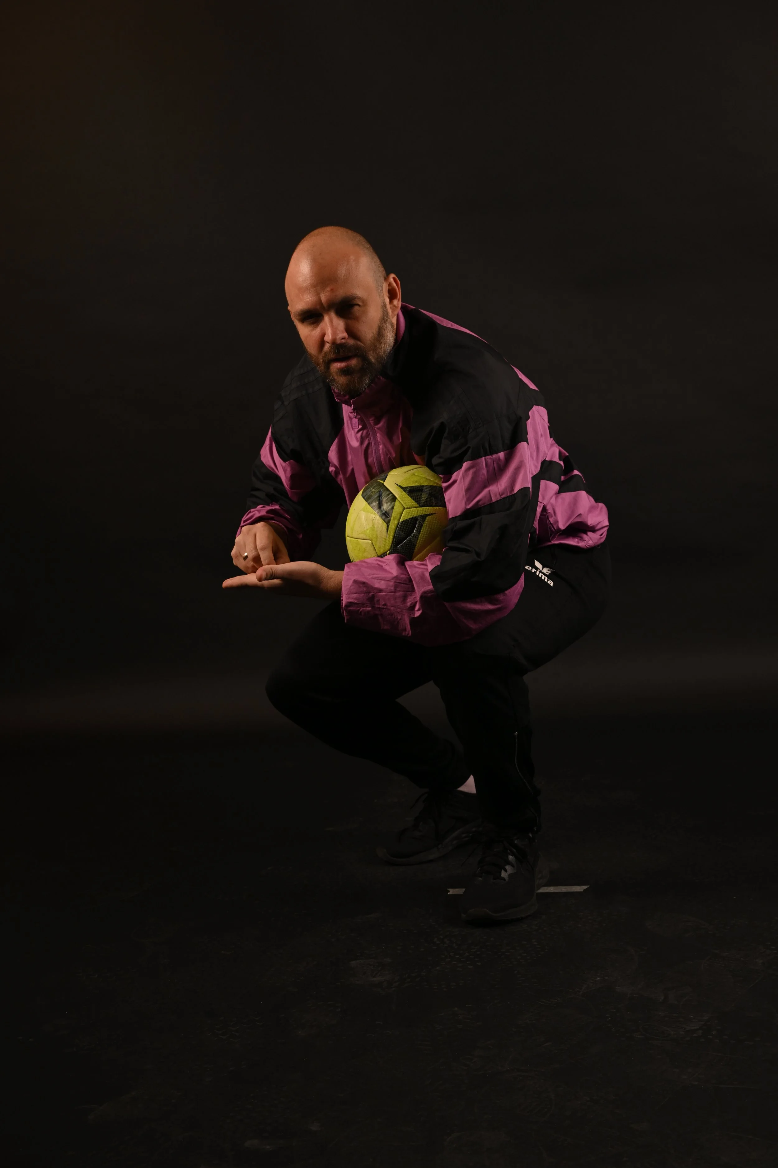 A man with a shaved head and beard wearing a black and pink sports jacket; he is squatting and holding a yellow and black soccer ball against a dark background.