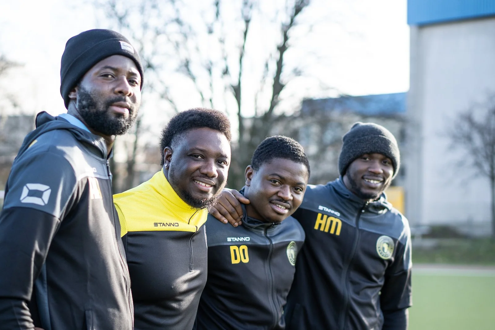 Four men standing close together outdoors, smiling, wearing black and yellow sports jackets, with trees and a building in the background.