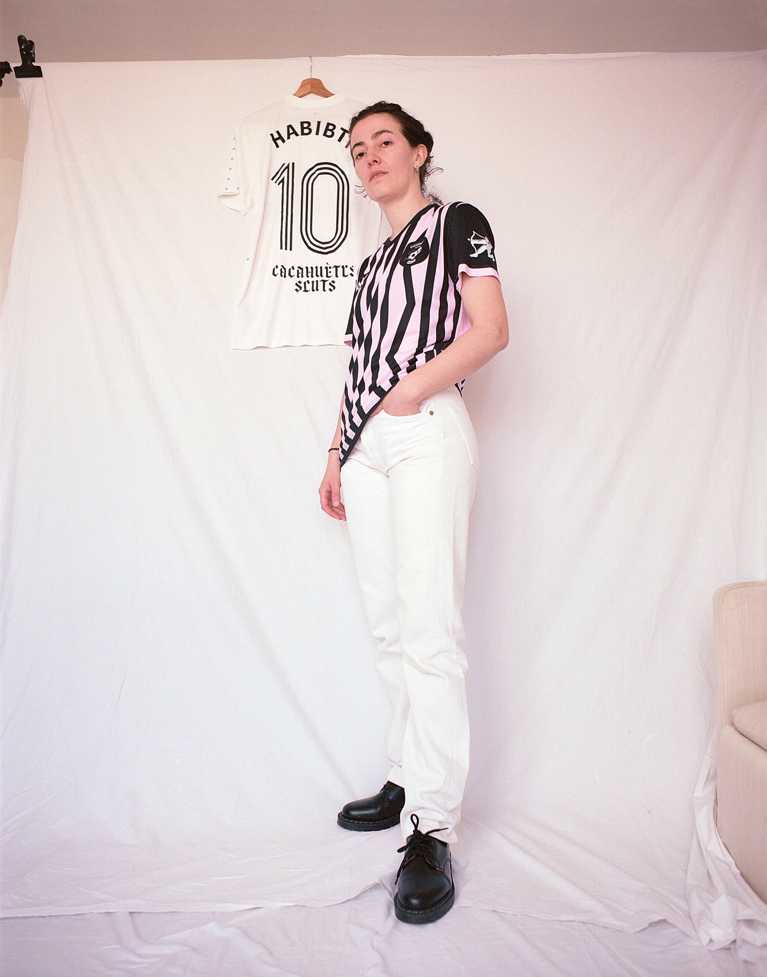 A woman with short dark hair and pierced ears wearing a red T-shirt with a white circle and the number 90 inside. She has a serious expression and is standing against a plain gray background.