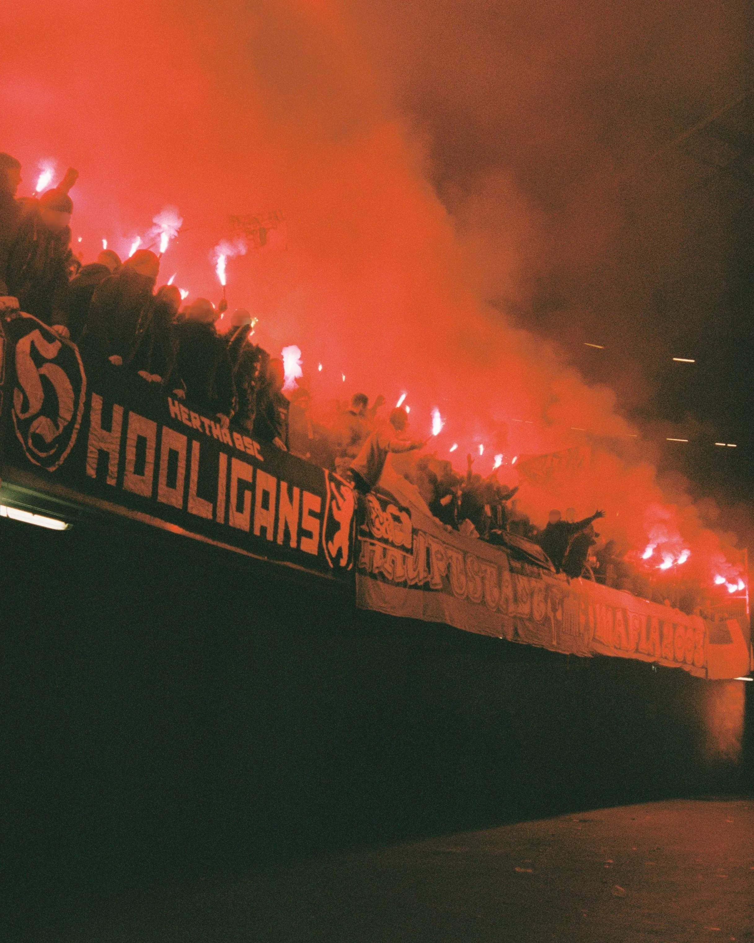 A group of people wearing helmets, standing on a barrier holding flares that emit bright light and smoke, with a banner on the barrier. The scene appears to be at night and may be a sports event or protest.