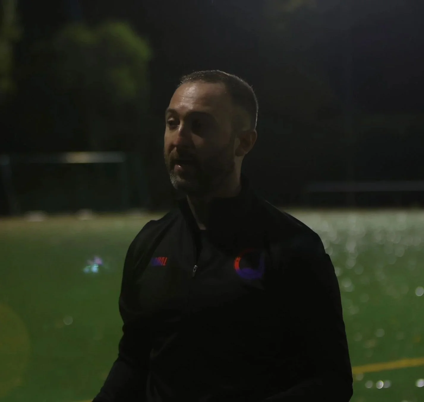 A man with short hair and a beard stands outdoors at night near what appears to be a sports field, with green turf and water reflections in the background, wearing a black jacket with logos.