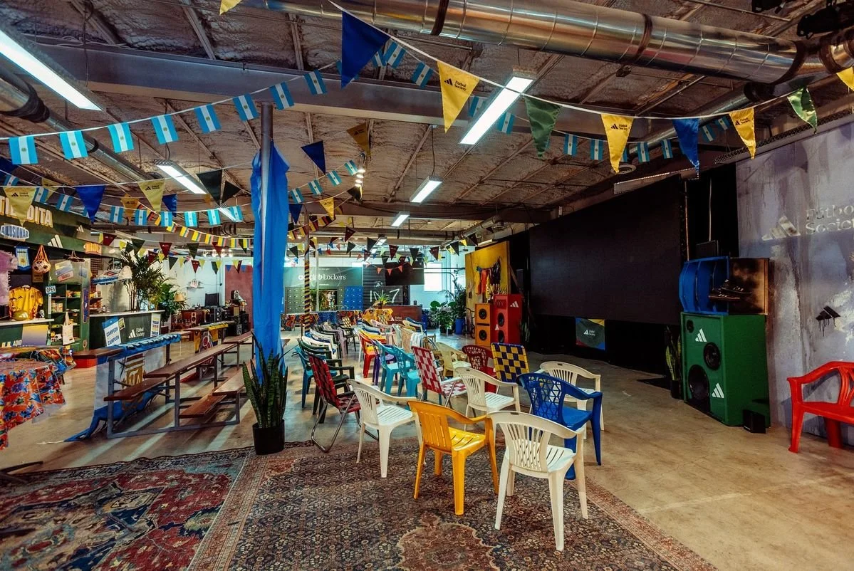 An indoor event space decorated with colorful flags and banners, featuring rows of chairs of various colors, a large screen, speakers, and a small stage area, with a jumbotron style screen in the background.
