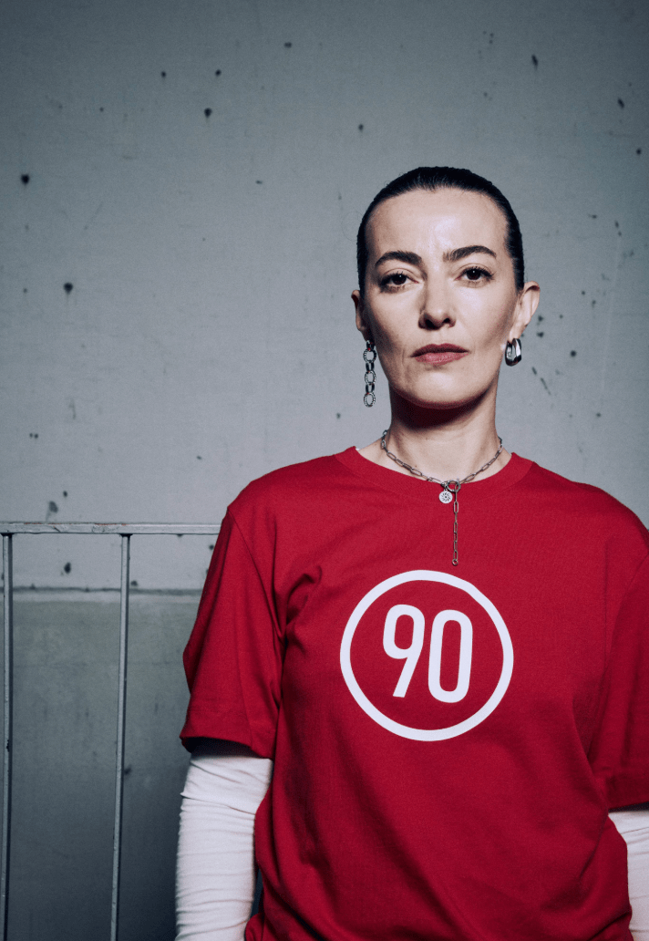 A woman with short dark hair and pierced ears wearing a red T-shirt with a white circle and the number 90 inside. She has a serious expression and is standing against a plain gray background.