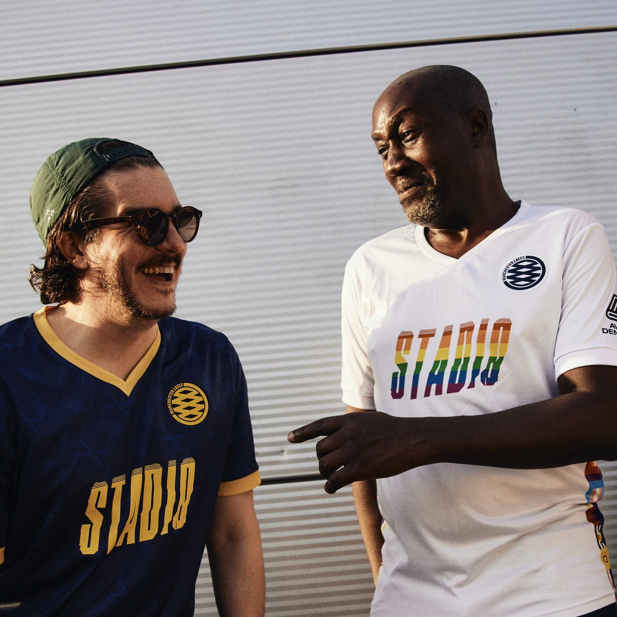 Two men wearing Manchester City jerseys with the word "STADIQ" in rainbow colors are smiling and talking outdoors in front of a metallic wall.