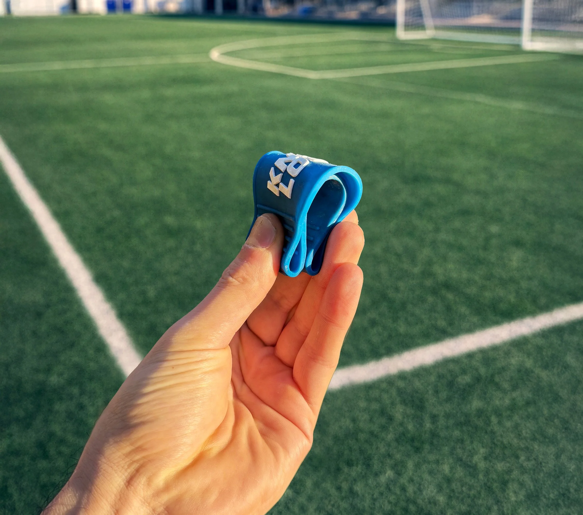 A person holding a blue silicone wristband from KnotLock on a soccer field with green artificial turf and white boundary lines.