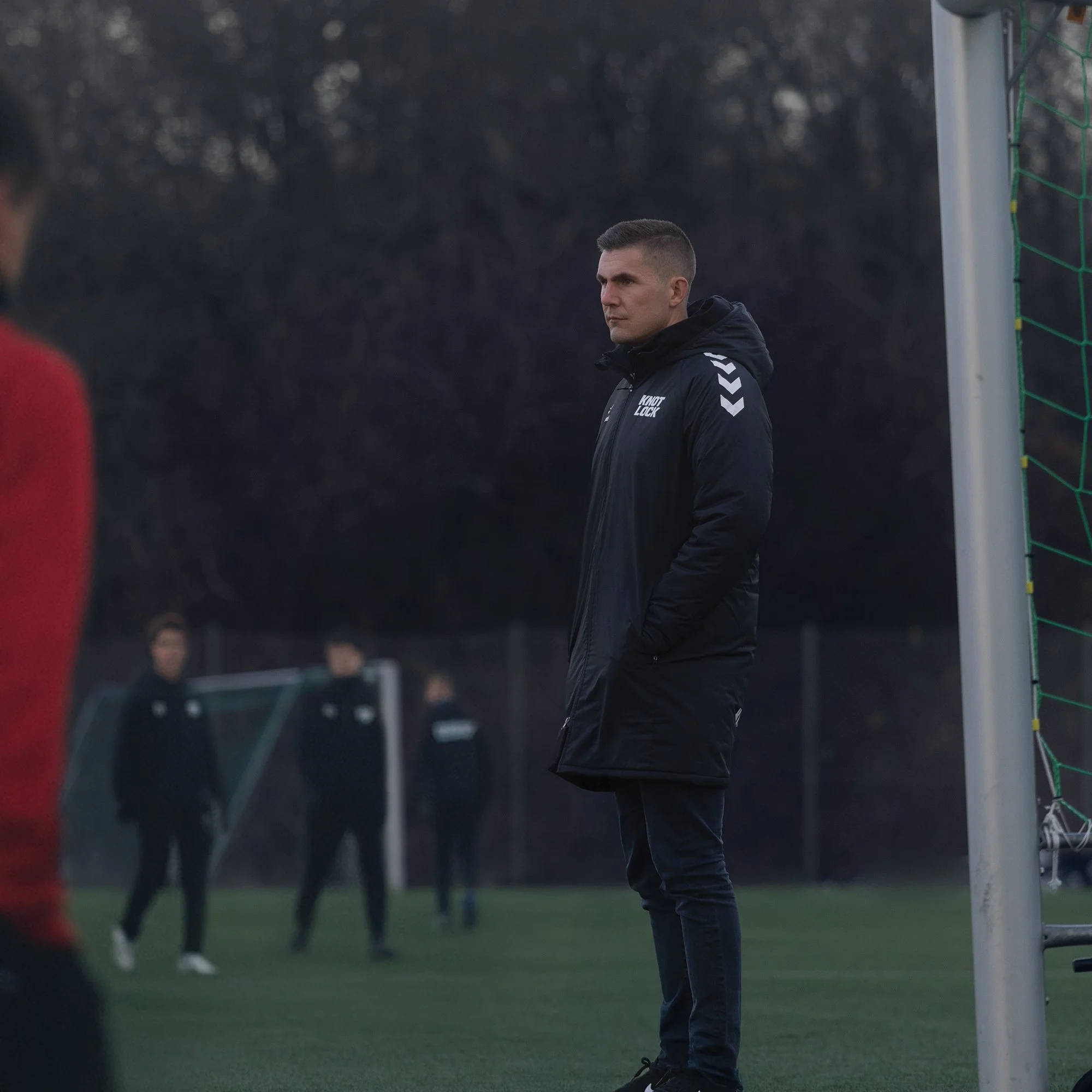 A man in a black coat and dark pants standing on a soccer field near the goalpost, observing others in the background.