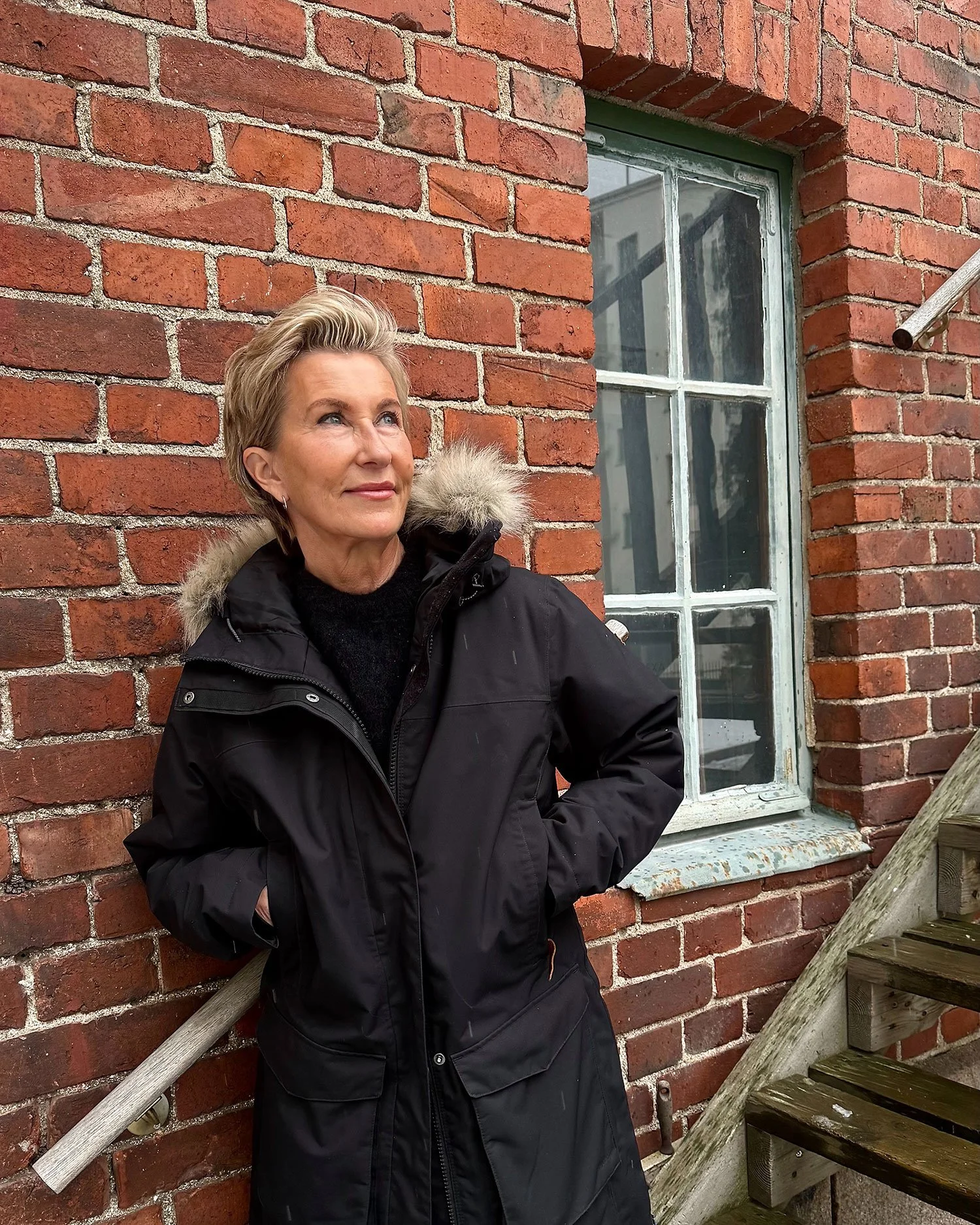 Leena Periaho seisoo punaisen tiilirakennuksen seinää vasten.