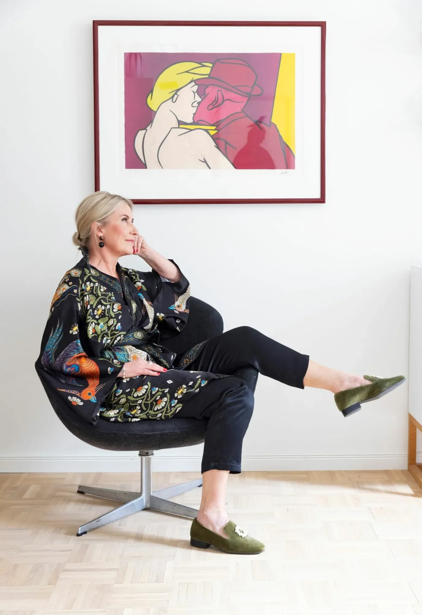 Nainen istuu nojatuolissa jalat ristittyinä katsoen sivulle. Taustalla on valkoinen seinä ja taidejuliste.