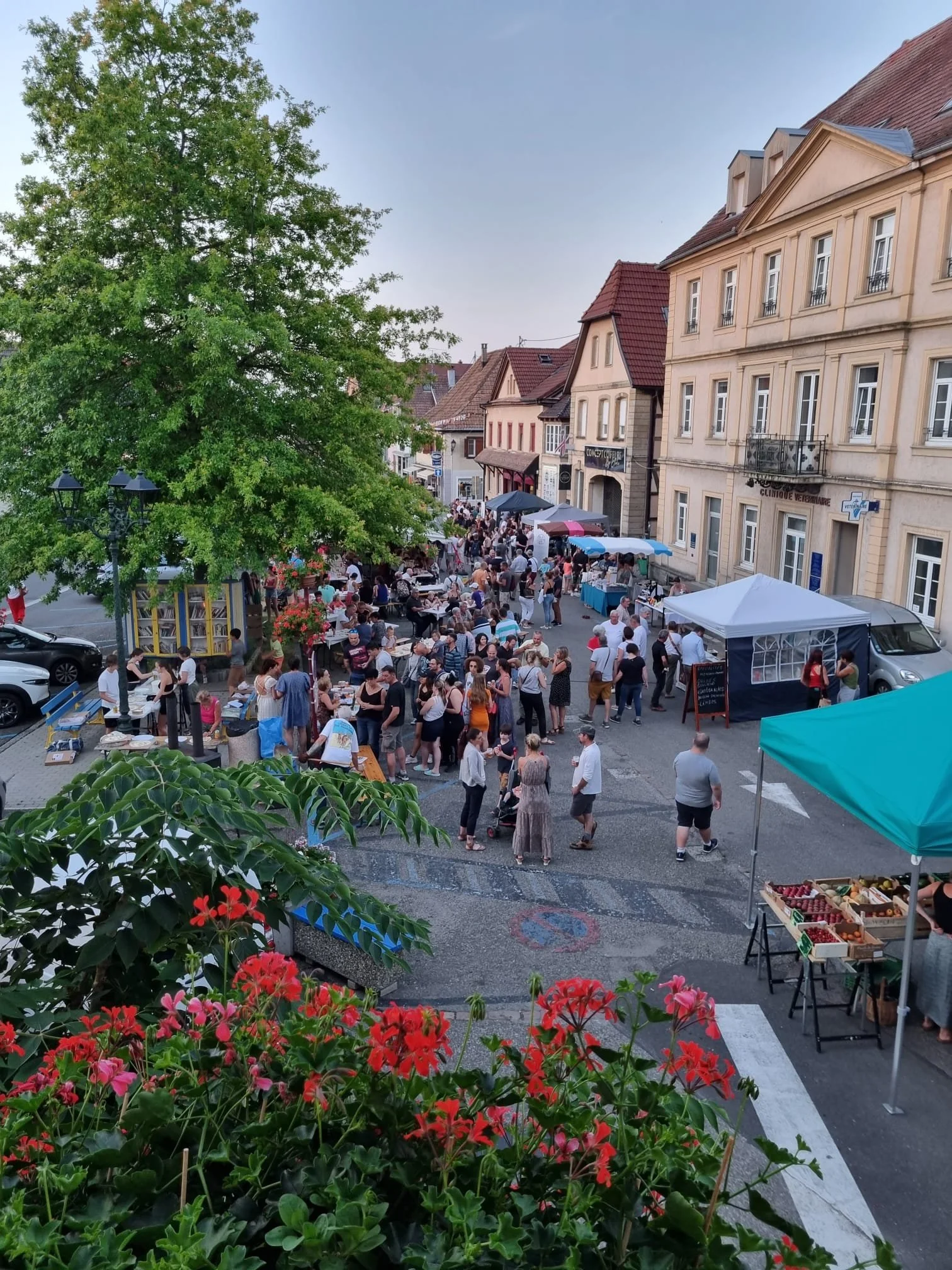 Marché en plein air avec des étals, des vendeurs et des acheteurs dans une rue bordée de bâtiments anciens, avec un arbre vert et des fleurs rouges en premier plan.
