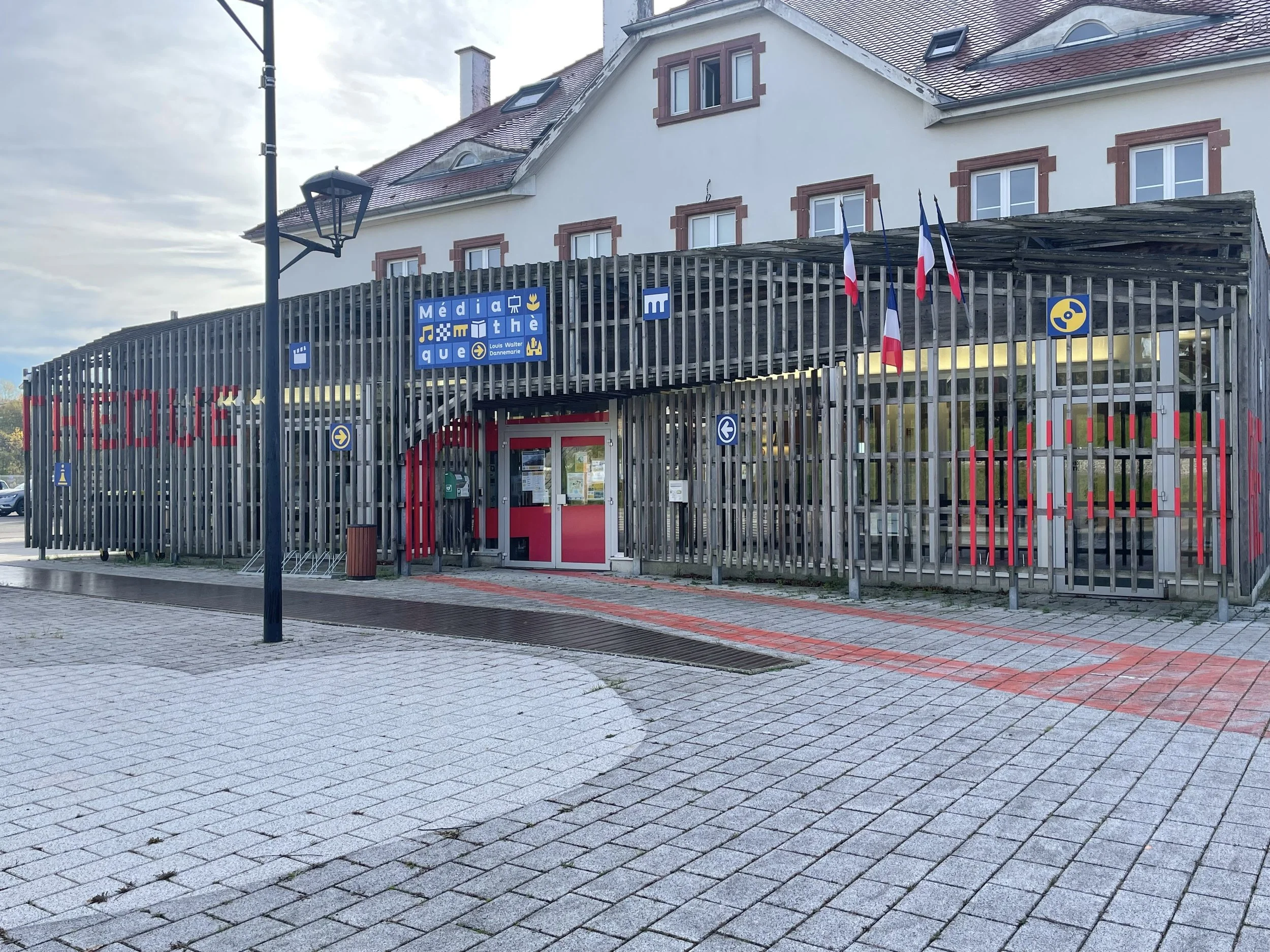 Le bâtiment est une bibliothèque médiathèque avec une façade en bois, drapeaux français, panneau bleus, un chemin en pierre devant l'entrée, et un lampadaire