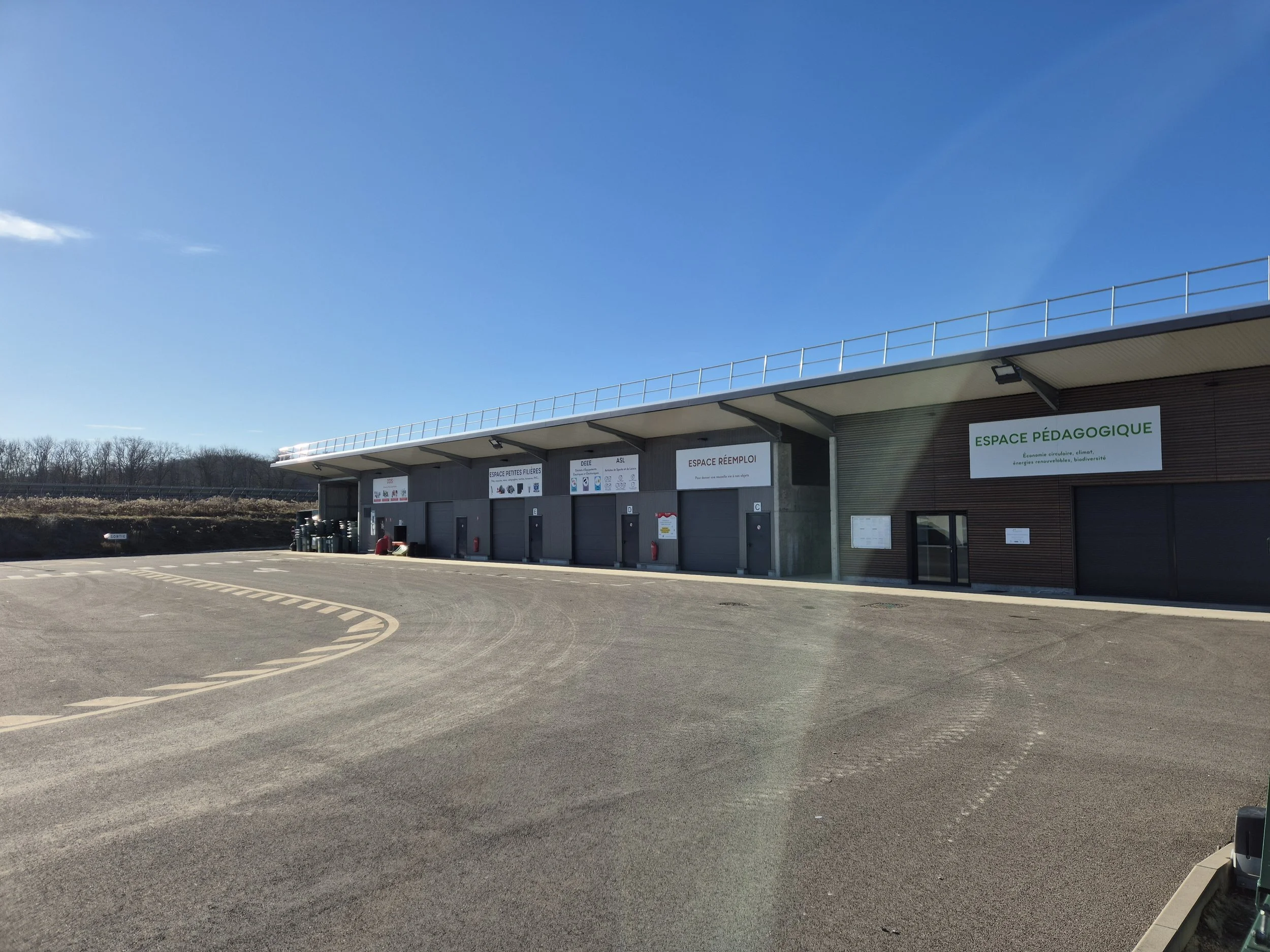 Bâtiment avec plusieurs panneaux et portes, espace pédagogique indiqué sur un panneau vert, ciel bleu clair, zone pavée sans véhicules.