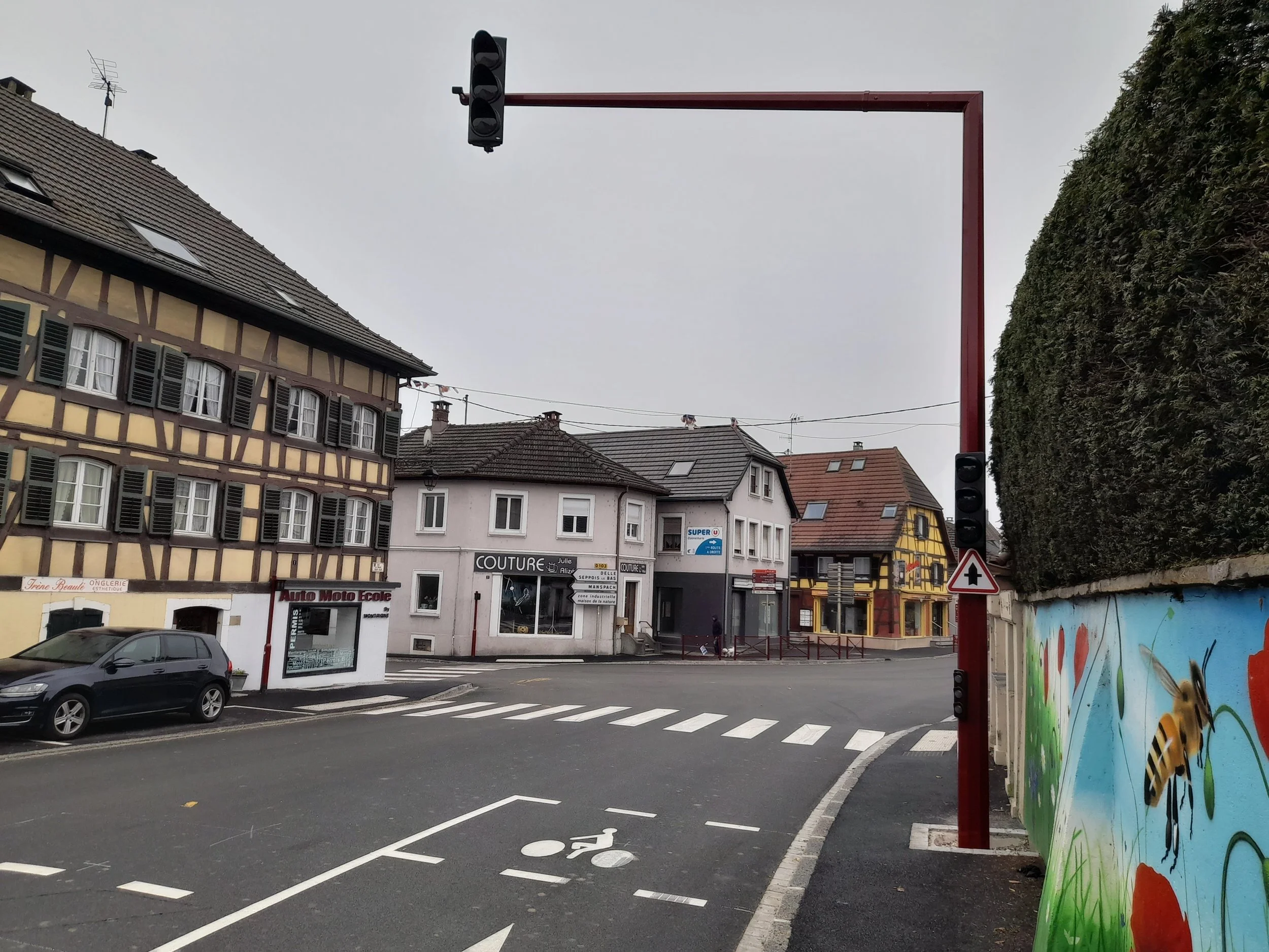 Une rue urbaine avec des bâtiments à façades en bois et une intersection avec un feu de signalisation rouge. Mur peint avec un dessin d'abeille et de fleurs sur le côté droit.