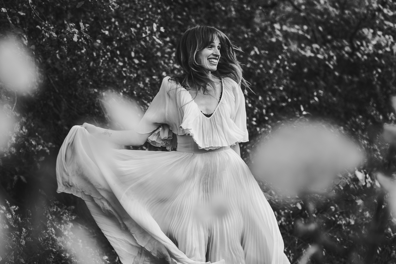 A woman in a flowing dress smiling, outdoors with trees in the background, in black and white.