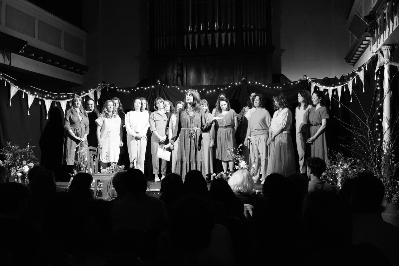 A group of women on stage participating in a performance or presentation, with some holding papers, decorated with string lights, bunting, and flowers, while an audience watches.