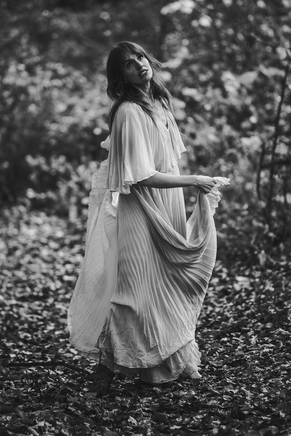 A woman in a flowing, pleated dress standing outdoors in a woodland area with fallen leaves on the ground. The photo is in black and white.
