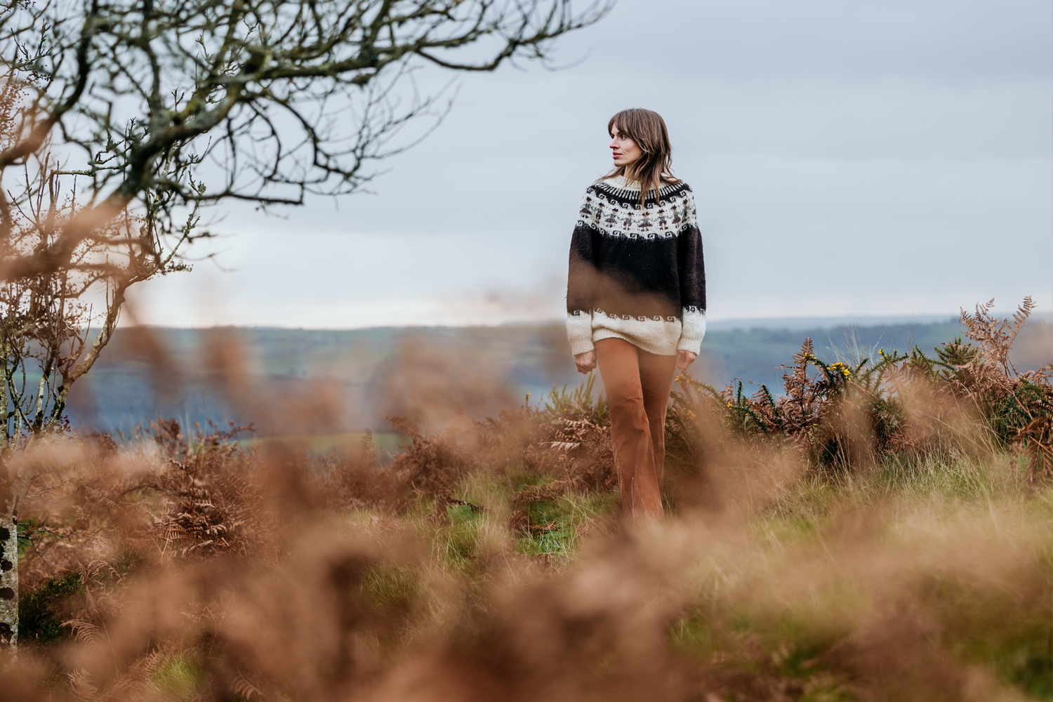 Woman standing outdoors in a rural landscape, wearing a black and white patterned sweater and brown pants, with blurred ferns in the foreground and rolling hills in the background on a cloudy day.