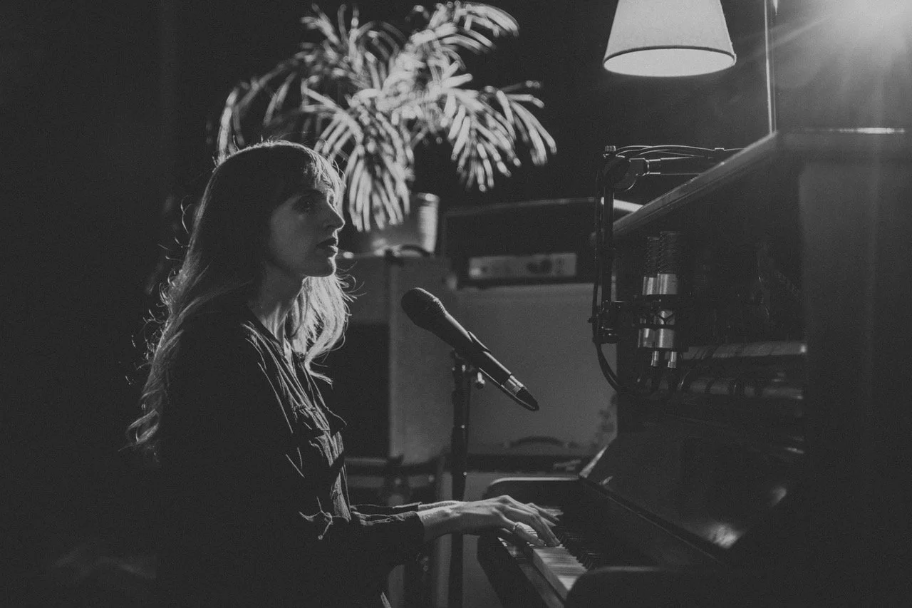 A woman playing a piano with a microphone in front of her, illuminated by a hanging lamp, in a dimly lit room with a large plant in the background.