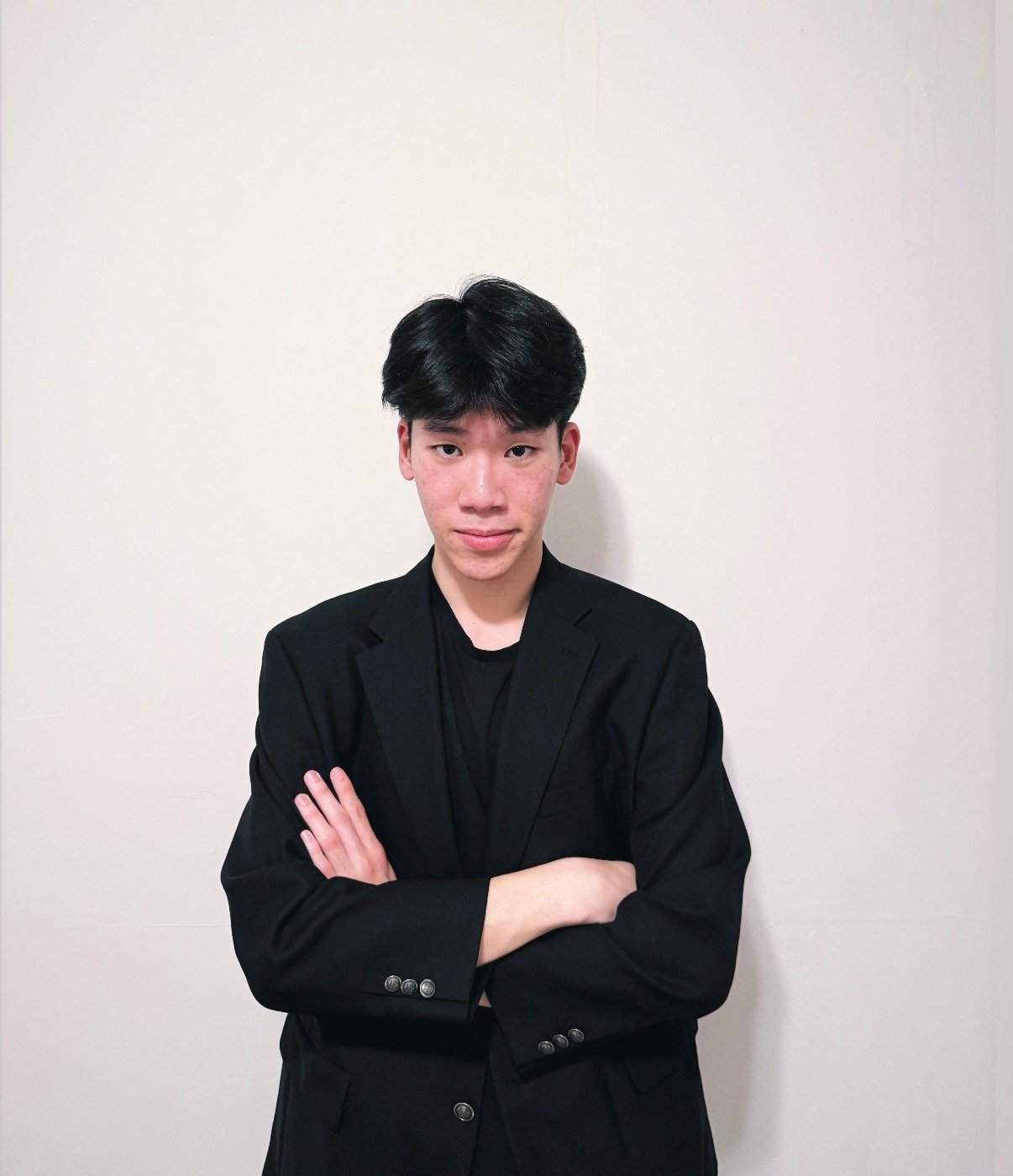 Young man with black hair wearing a black blazer and T-shirt, standing with arms crossed in front of a plain white wall.