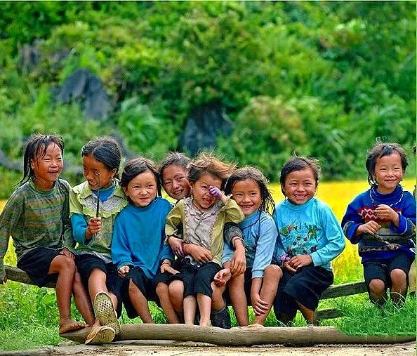 Group of children sitting outdoors on a log, smiling and laughing, with green foliage in the background.
