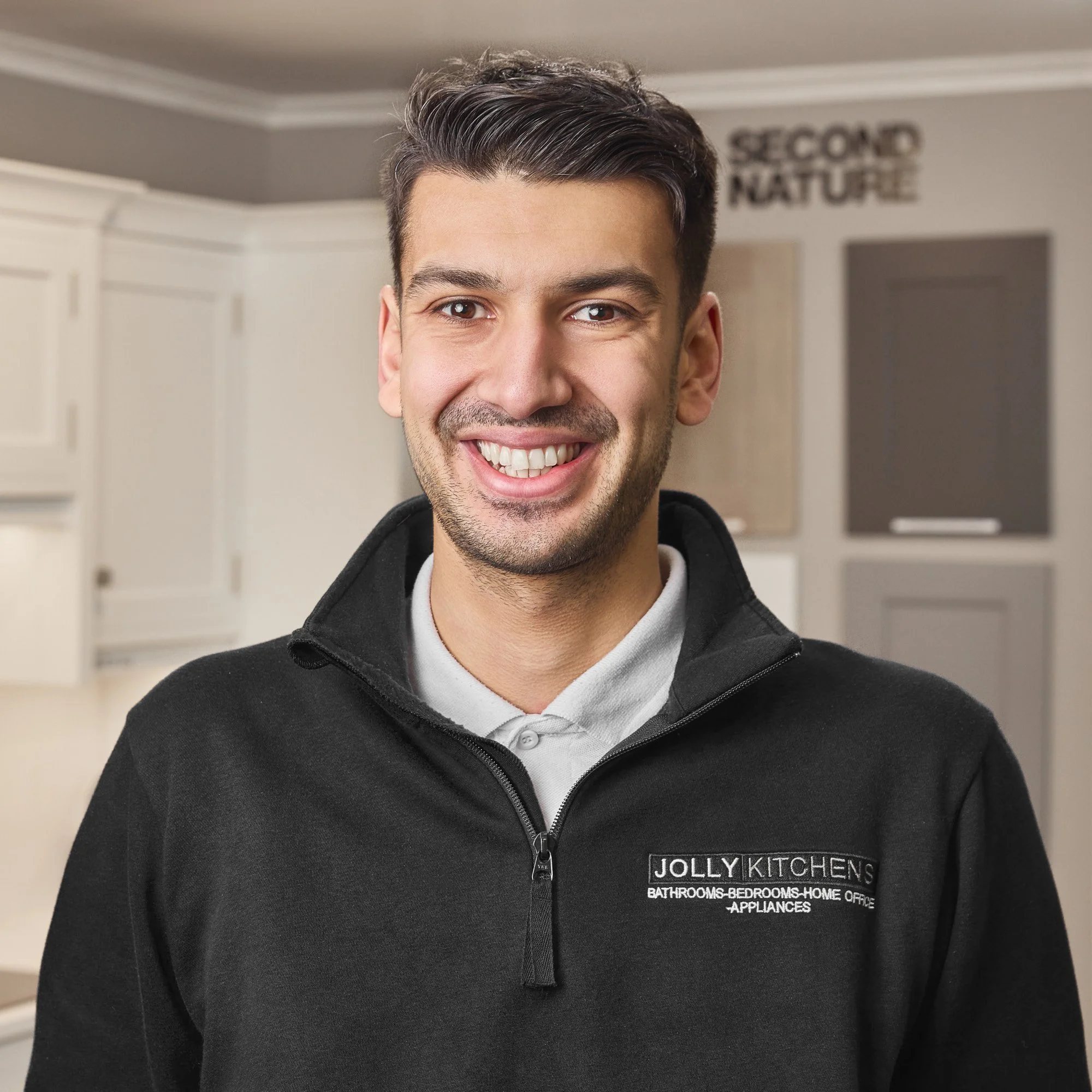 A smiling man wearing a Jolly Kitchens uniform standing in a kitchen with white cabinets