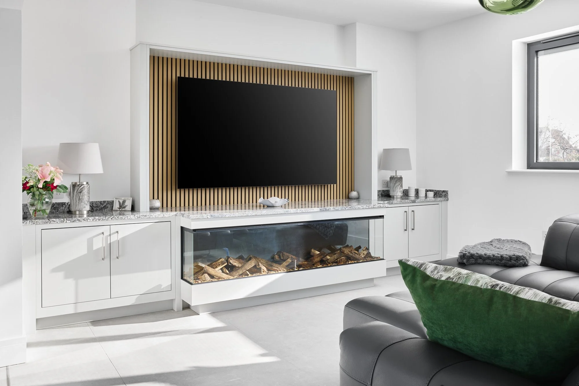 Modern living room with a built-in entertainment center featuring a large black flat-screen TV on a wooden slatted background, a fireplace with logs, white cabinetry, a gray sofa with a green pillow, and a window with a view outside.
