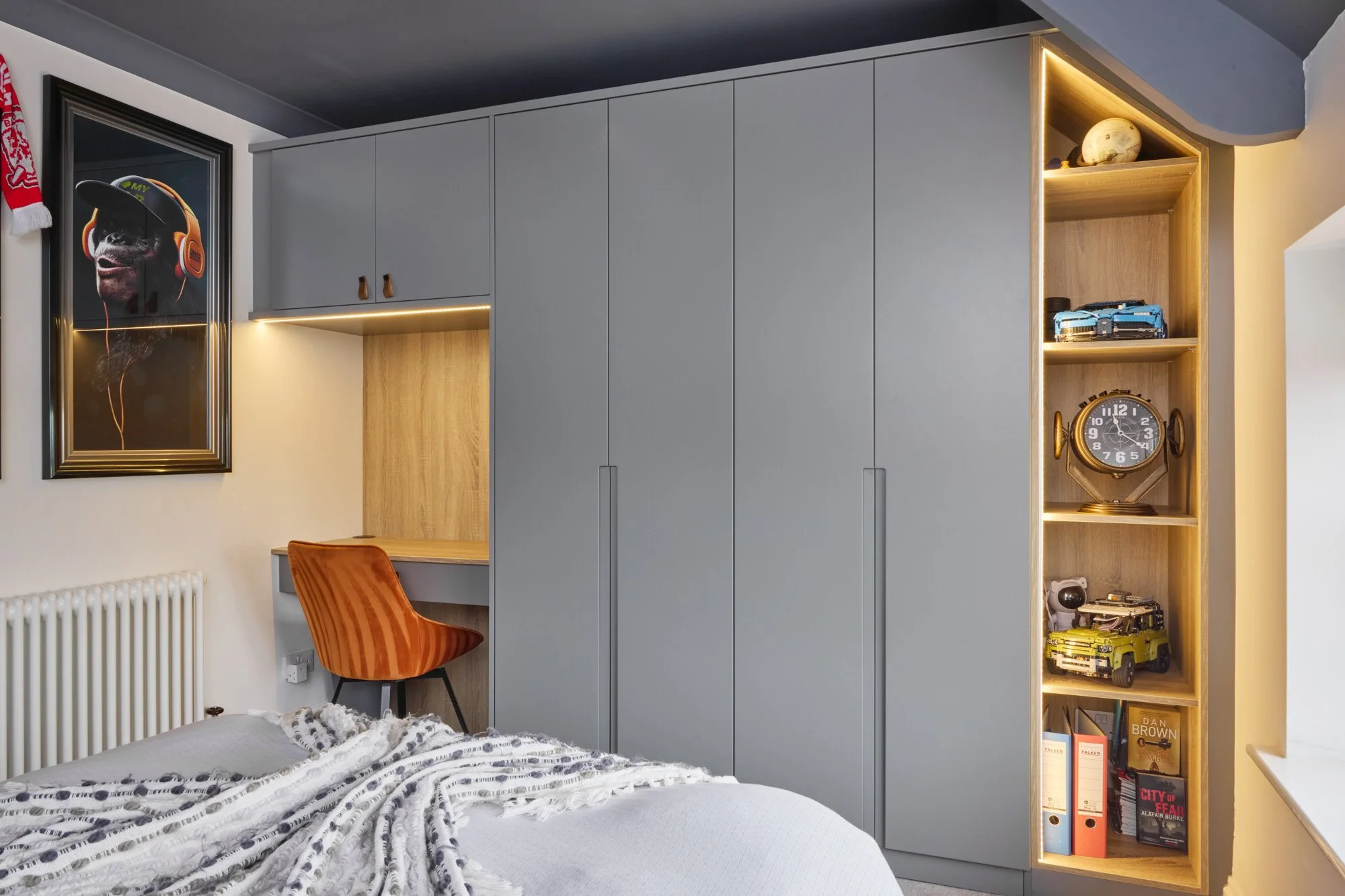 A bedroom corner with a gray wardrobe, a small built-in desk with a burnt orange chair, a framed dog picture wearing headphones, and a wooden shelving unit with toys and books.