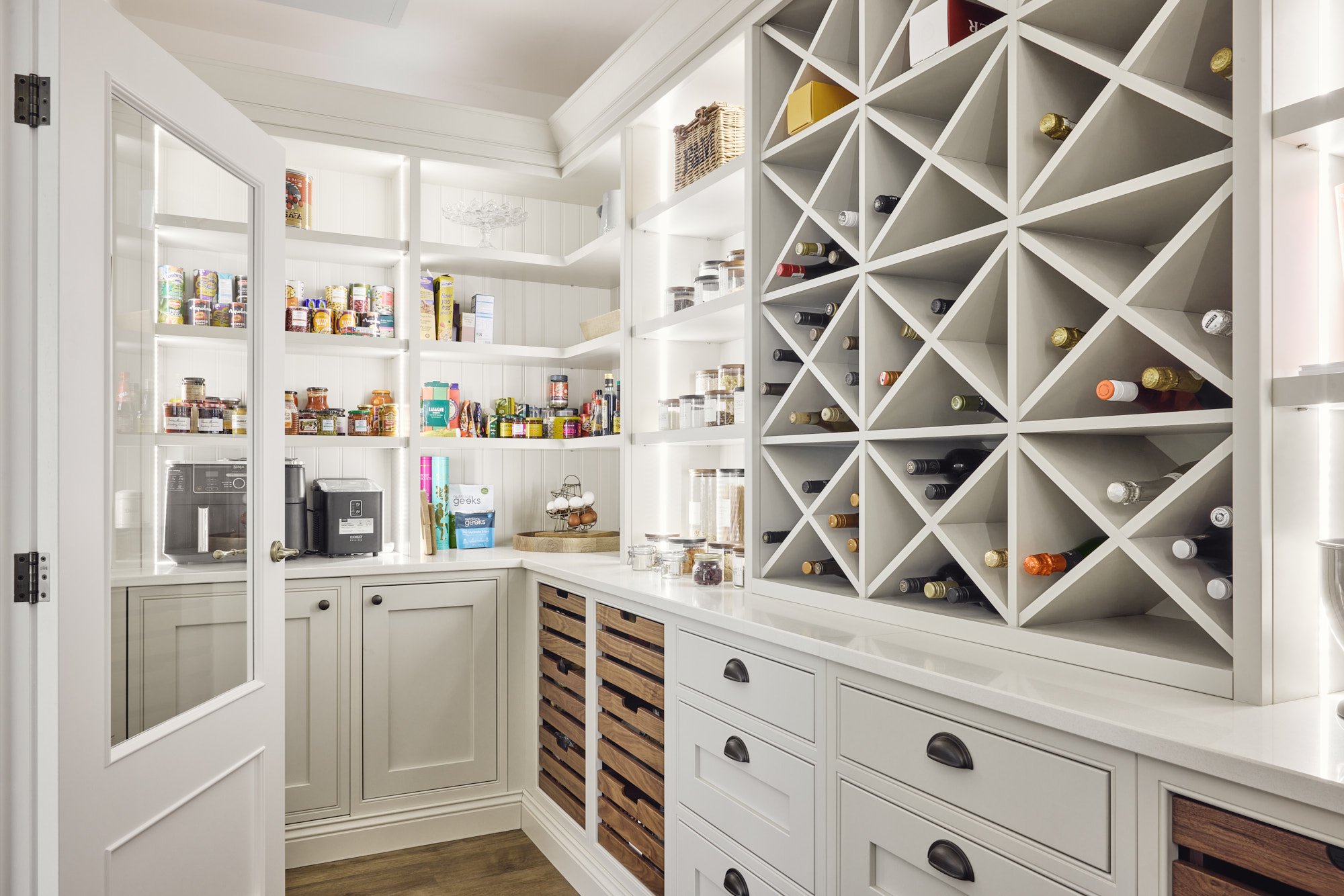 A well-organized pantry with white cabinetry, wine storage racks, open shelves, and various food items and kitchen appliances.