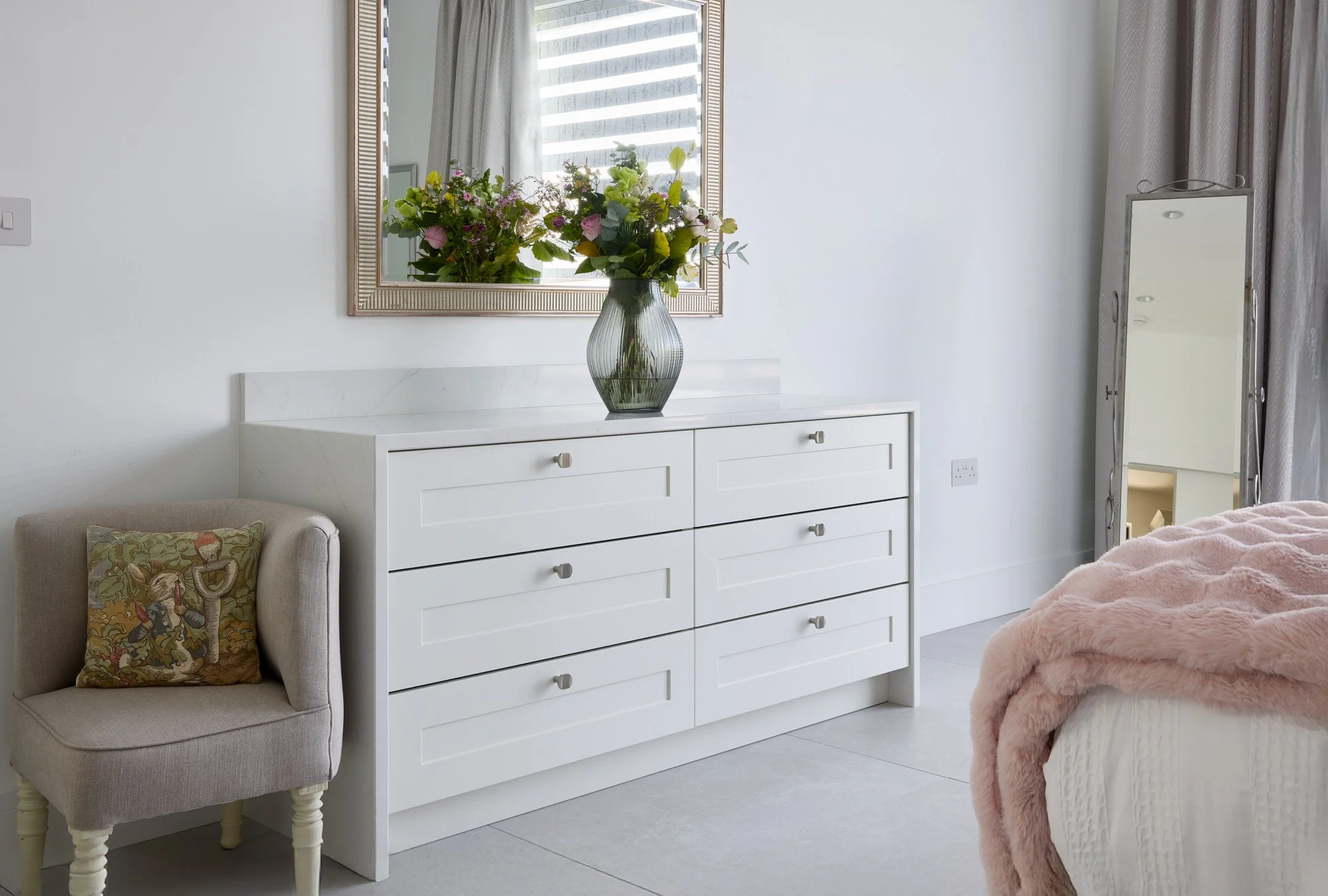 A white dresser with six drawers and a marble top, holding a large glass vase with a bouquet of mixed flowers. A small upholstered chair with a decorative pillow is beside the dresser. A rectangular mirror hangs above the dresser, and a tall, narrow mirror stands to the right, near a window with curtains. Part of a bed with a pink blanket is visible in the foreground.