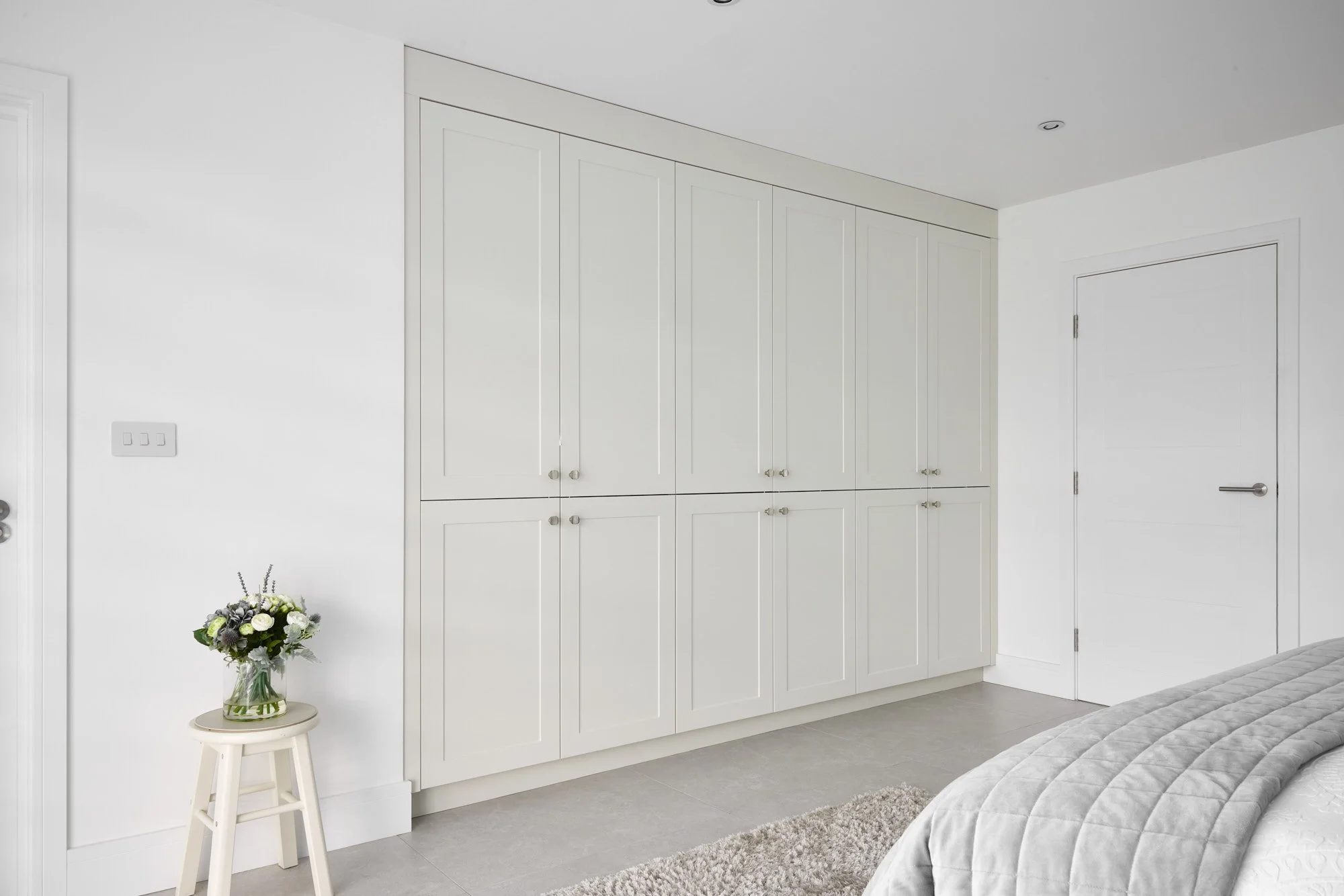 A bedroom with white walls, a large white wardrobe with multiple doors, a small stool with a flower vase, part of a bed with gray bedding, and a door with a silver handle.