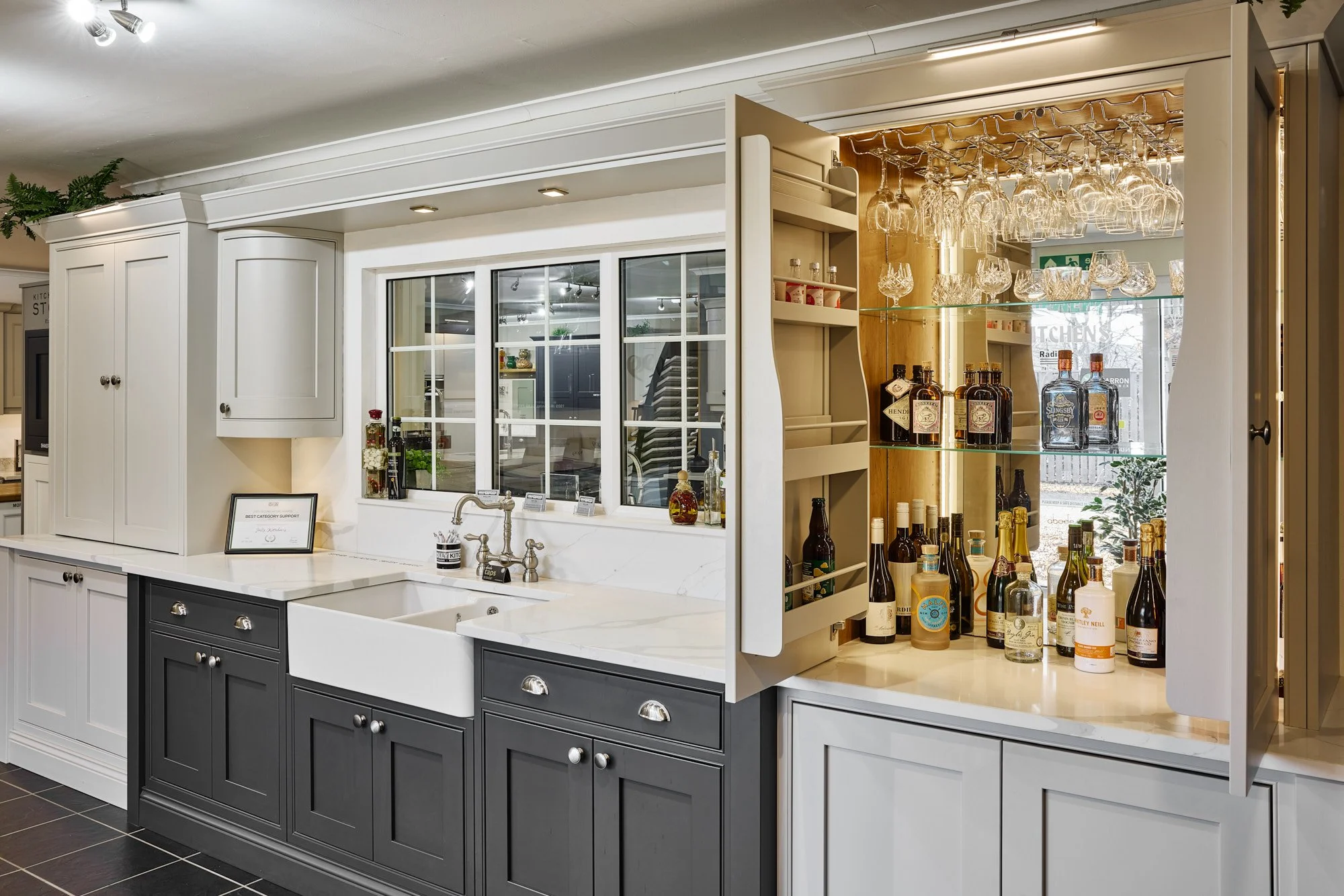 Kitchen bar area with glassware and bottles of liquor on display, white and dark gray cabinetry, and a window above the sink.