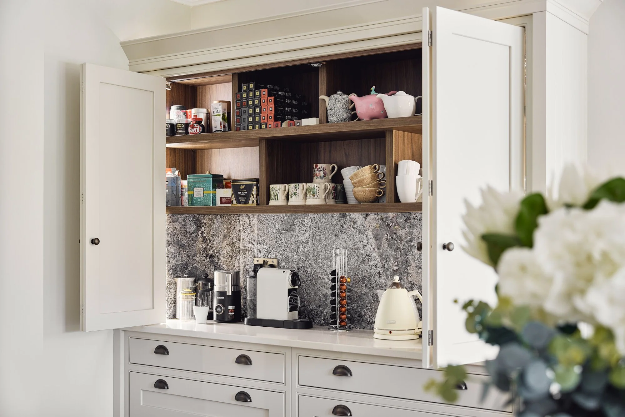 Open kitchen cabinet with coffee maker, kettle, and coffee capsules on the countertop, and cups, teapots, and storage boxes on the shelves.