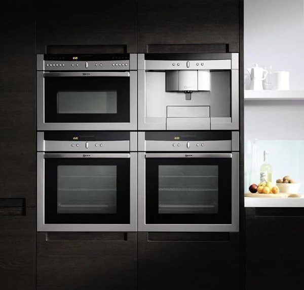 Four built-in stainless steel kitchen appliances: a microwave oven on top left, a coffee machine on top right, and two double electric ovens below, installed in black cabinetry with a white shelf holding a teapot, cups, and apples
