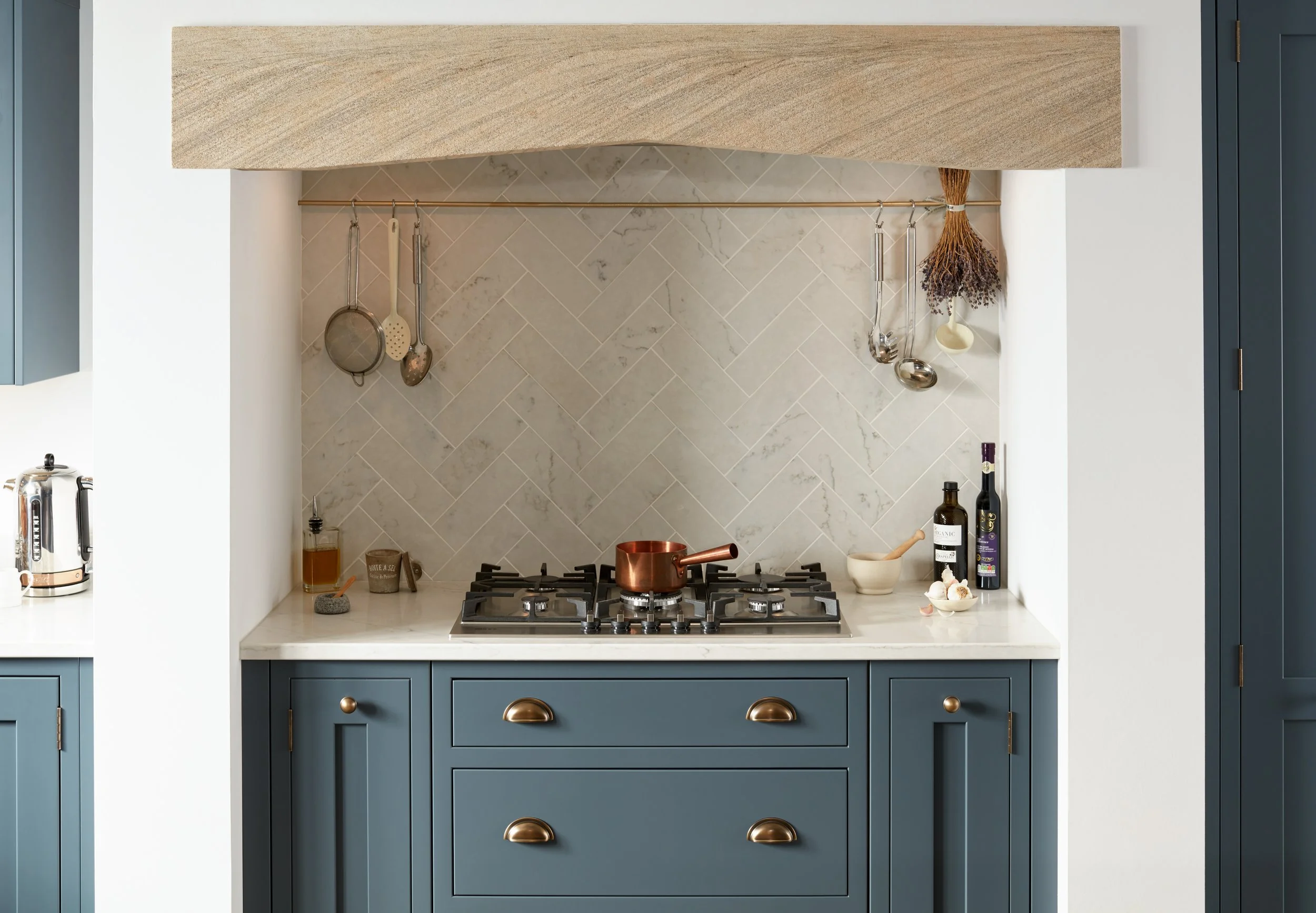 Kitchen with a blue cabinet, beige countertop, and a beige herringbone backsplash, featuring a copper saucepan on a gas stove and hanging utensils.