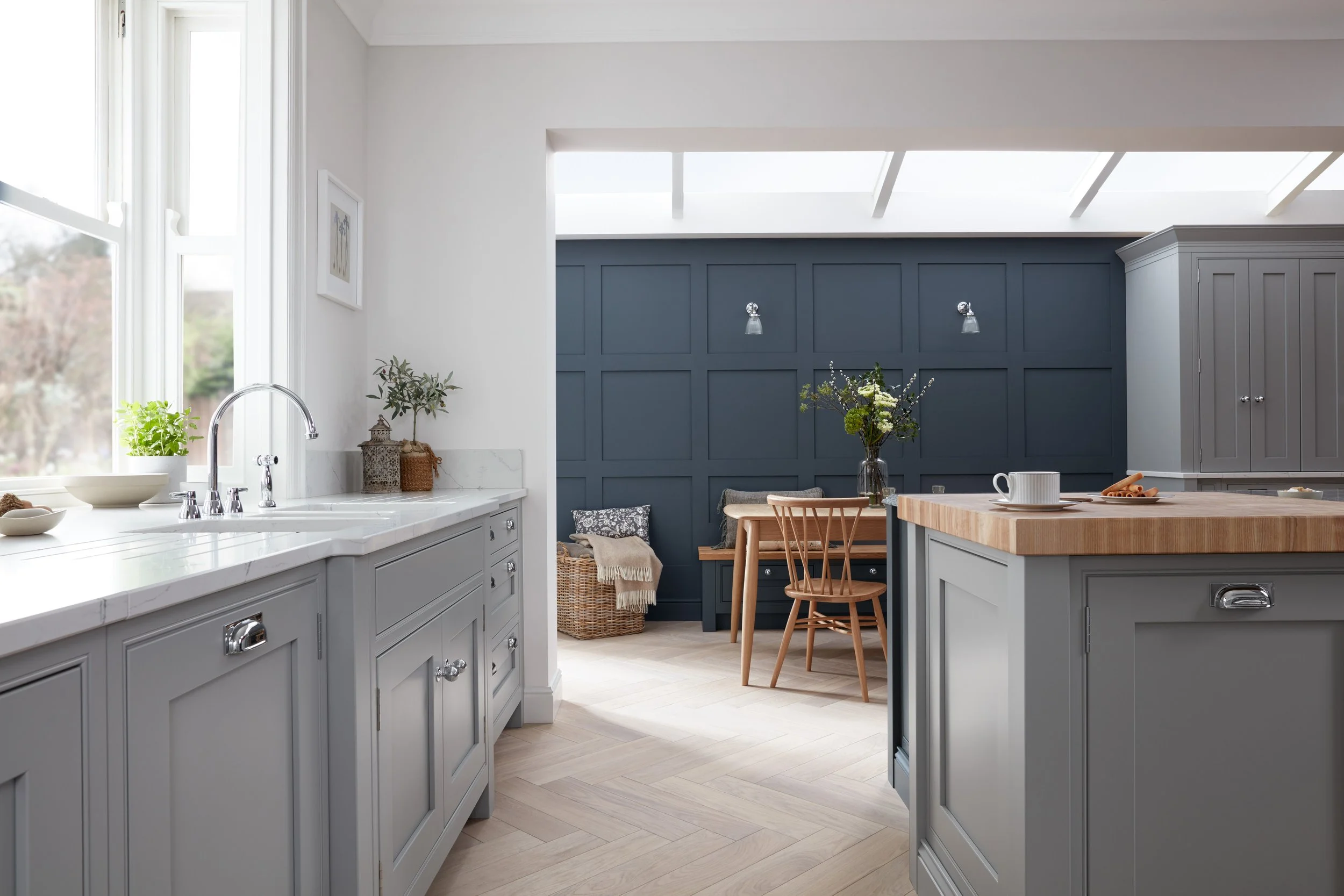 A bright modern kitchen with gray cabinetry, white countertops, and a central island. There is a window with natural light, a sink, and a small dining nook with a wooden table and chairs against a dark blue paneled wall, decorated with a floral arrangement.
