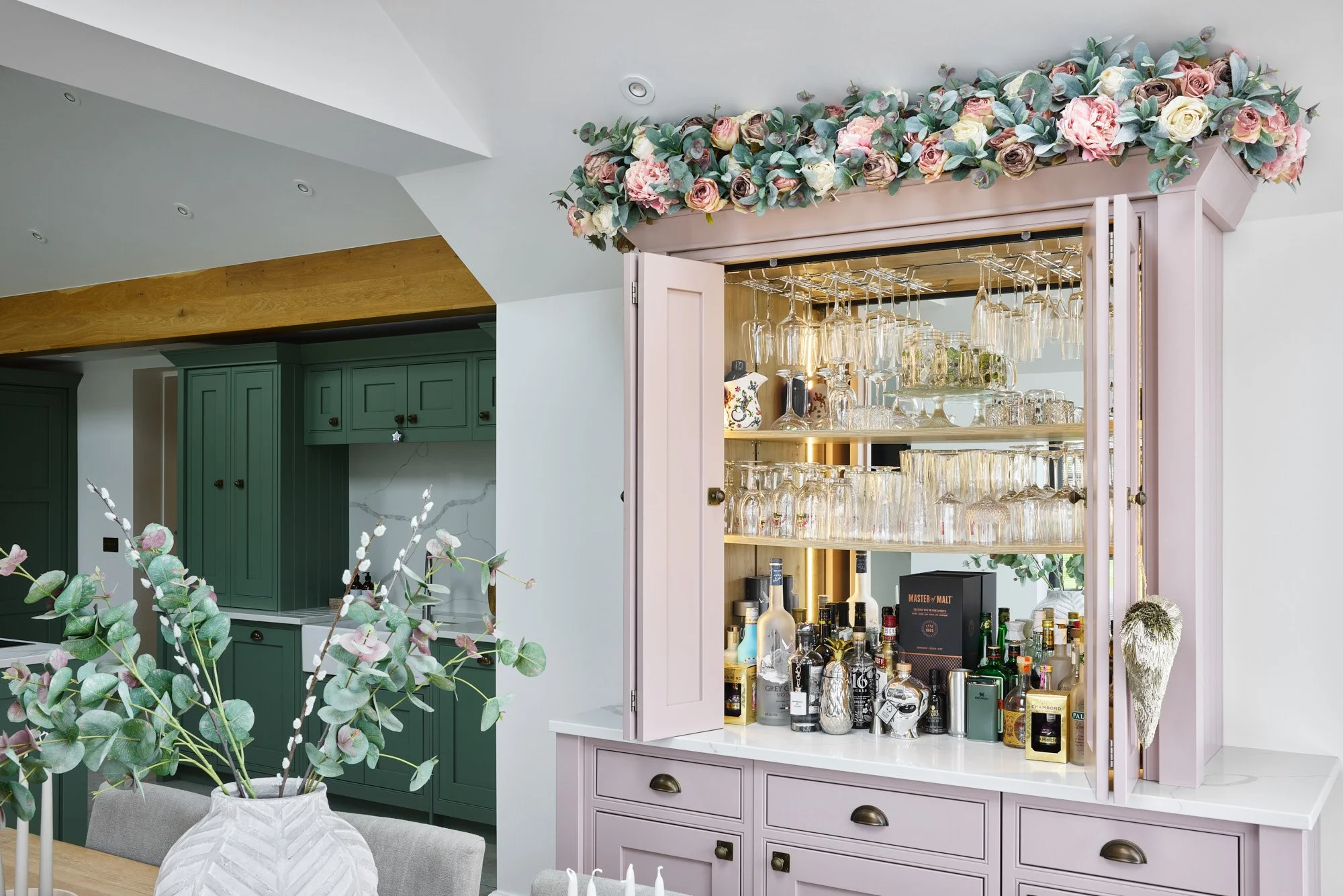 A pink bar cabinet decorated with a floral arrangement on top, filled with various bottles, glasses, and barware in a modern kitchen and dining area.