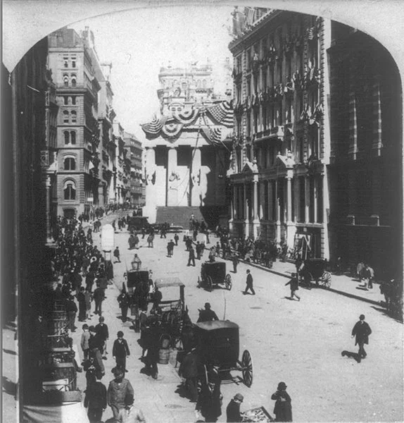 Historic city street scene with men walking and horse-drawn carriages, large buildings, and a decorated monument in the background.