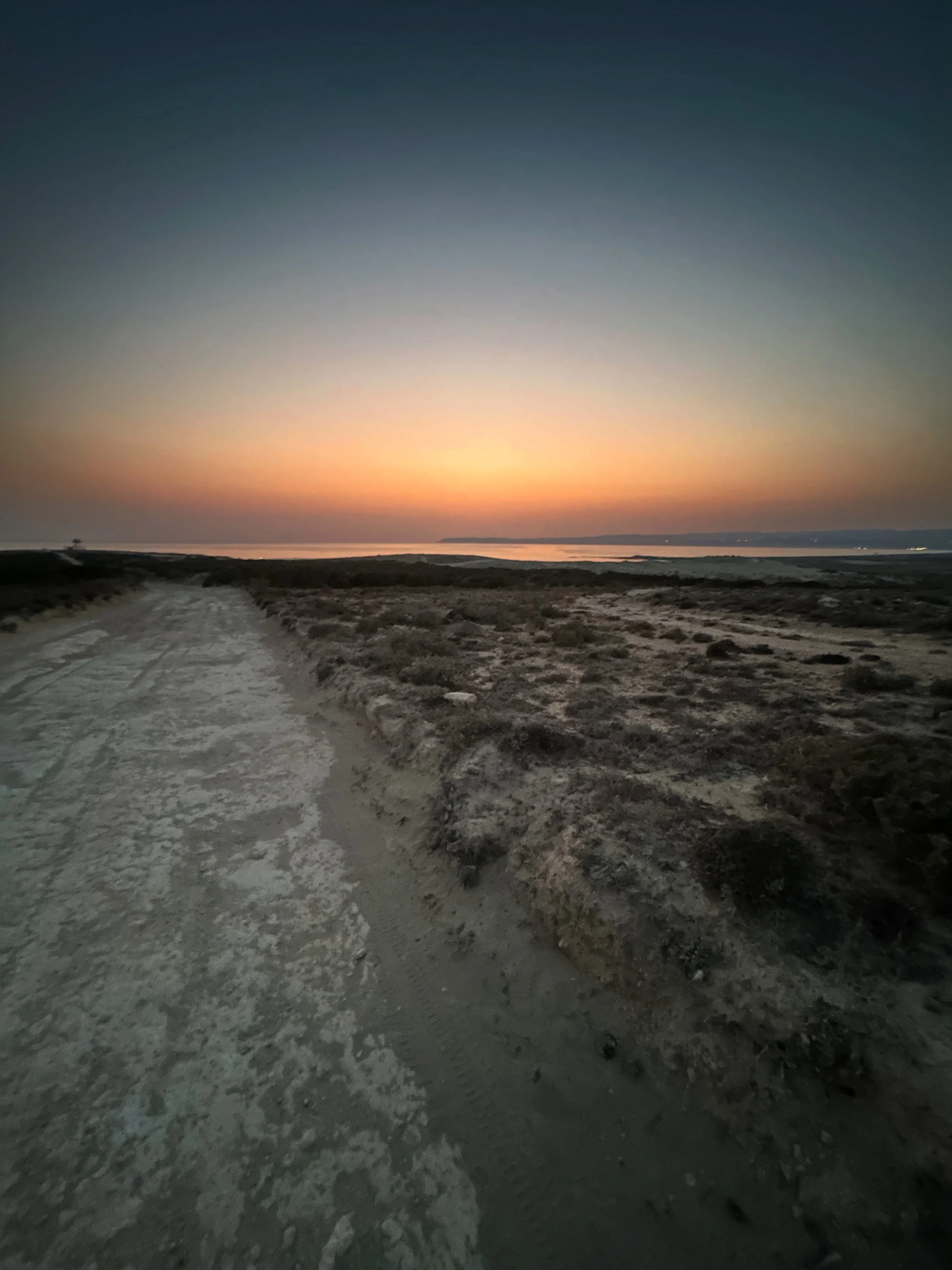 Dirt path in a coastal area during sunset or sunrise with a calm body of water and distant land in the background, and a gradient sky transitioning from dark to light.