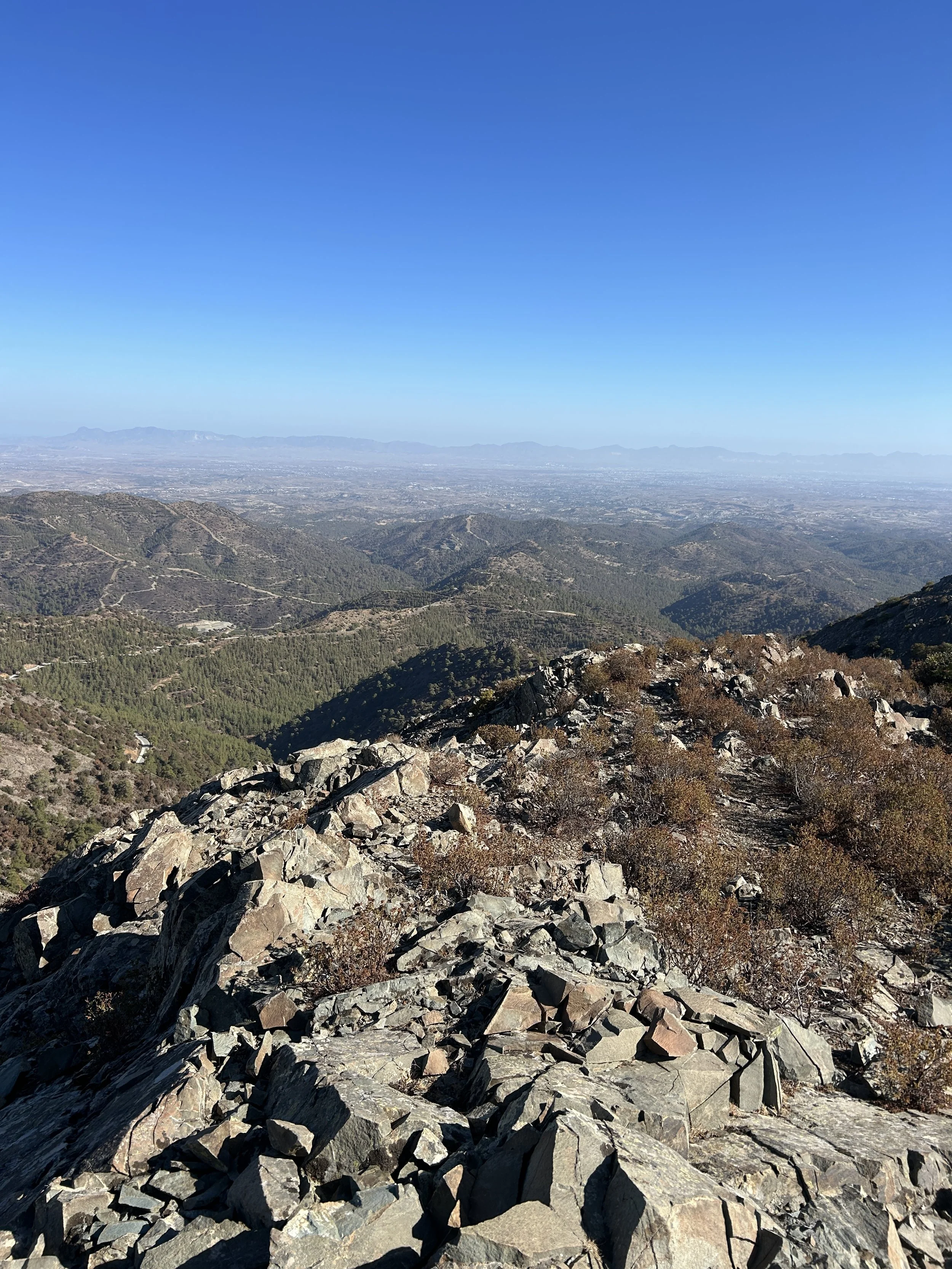 A mountainous landscape with rocky terrain in the foreground, green hills and valleys in the midground, and a clear blue sky overhead.