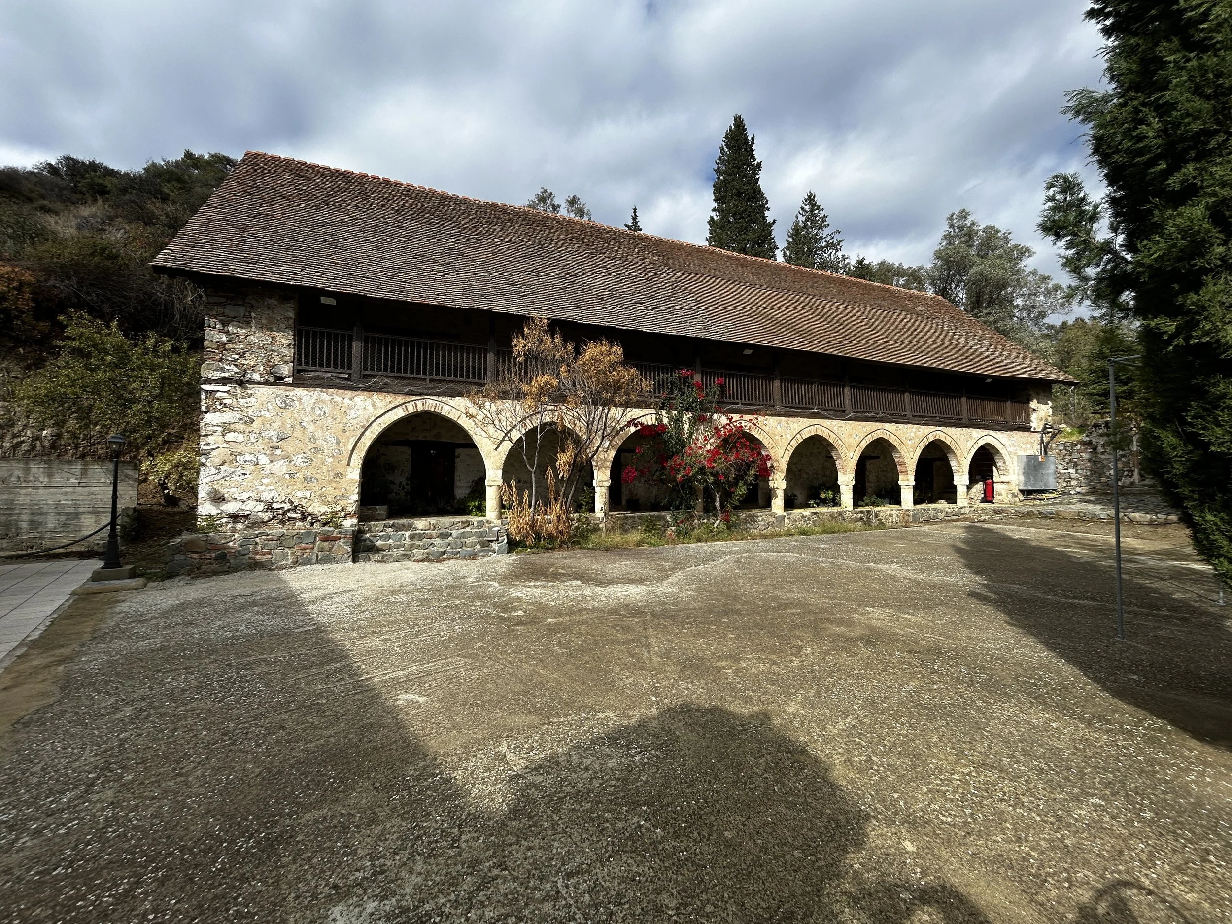 A historic stone building with a red tile roof and arched openings on the ground floor, surrounded by trees and bushes, under a cloudy sky.
